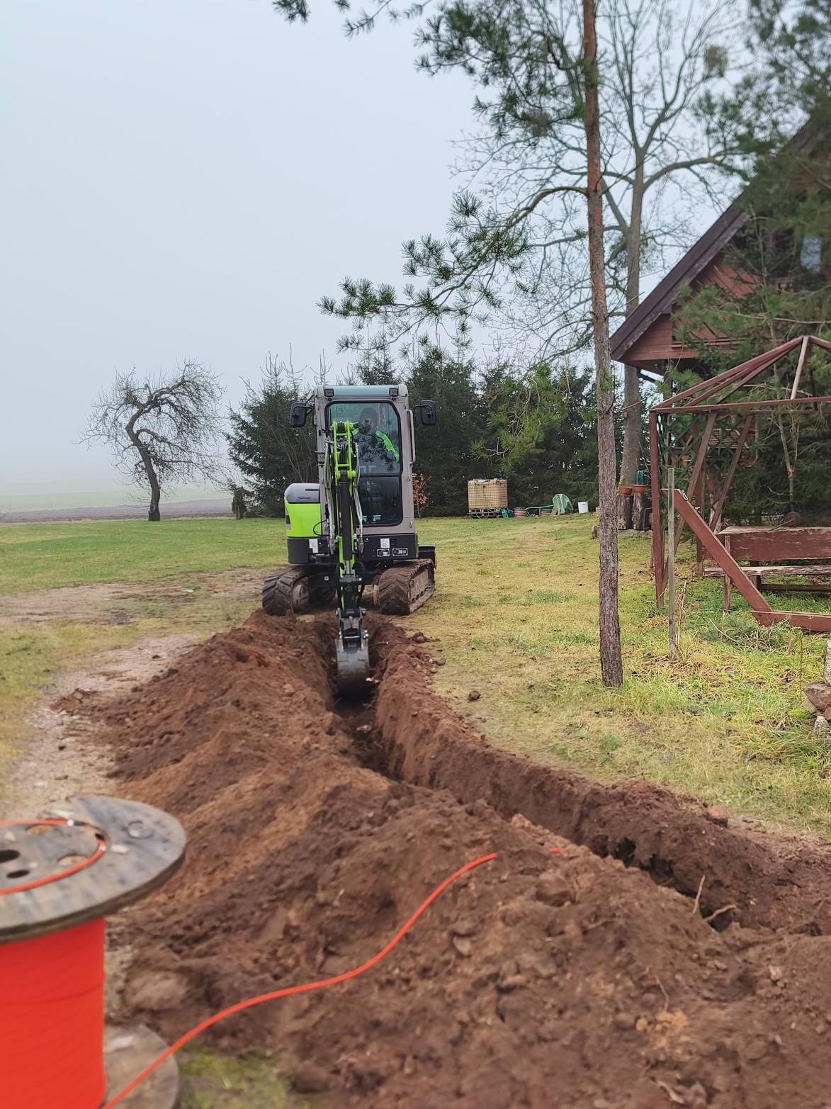 Mini koparka z operatorem kopie wąski wykop w ziemi na zewnątrz, obok leży zwój pomarańczowego kabla. W tle dom i drzewa.