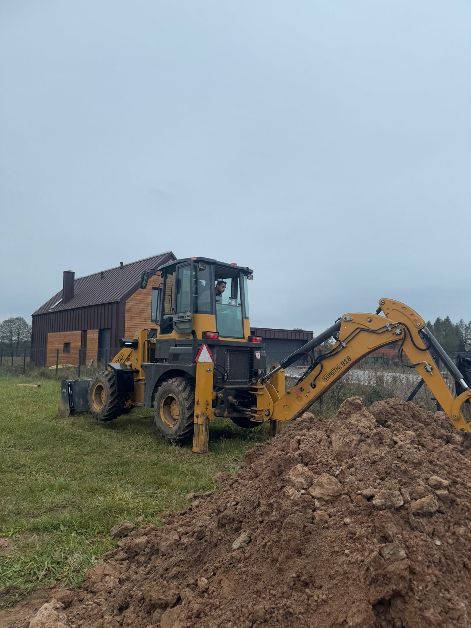Żółta koparko-ładowarka Günstig 918 na trawiastym terenie z pryzmą ziemi, w tle nowoczesny dom z brązową elewacją i szarym niebem. Operator w kabinie.