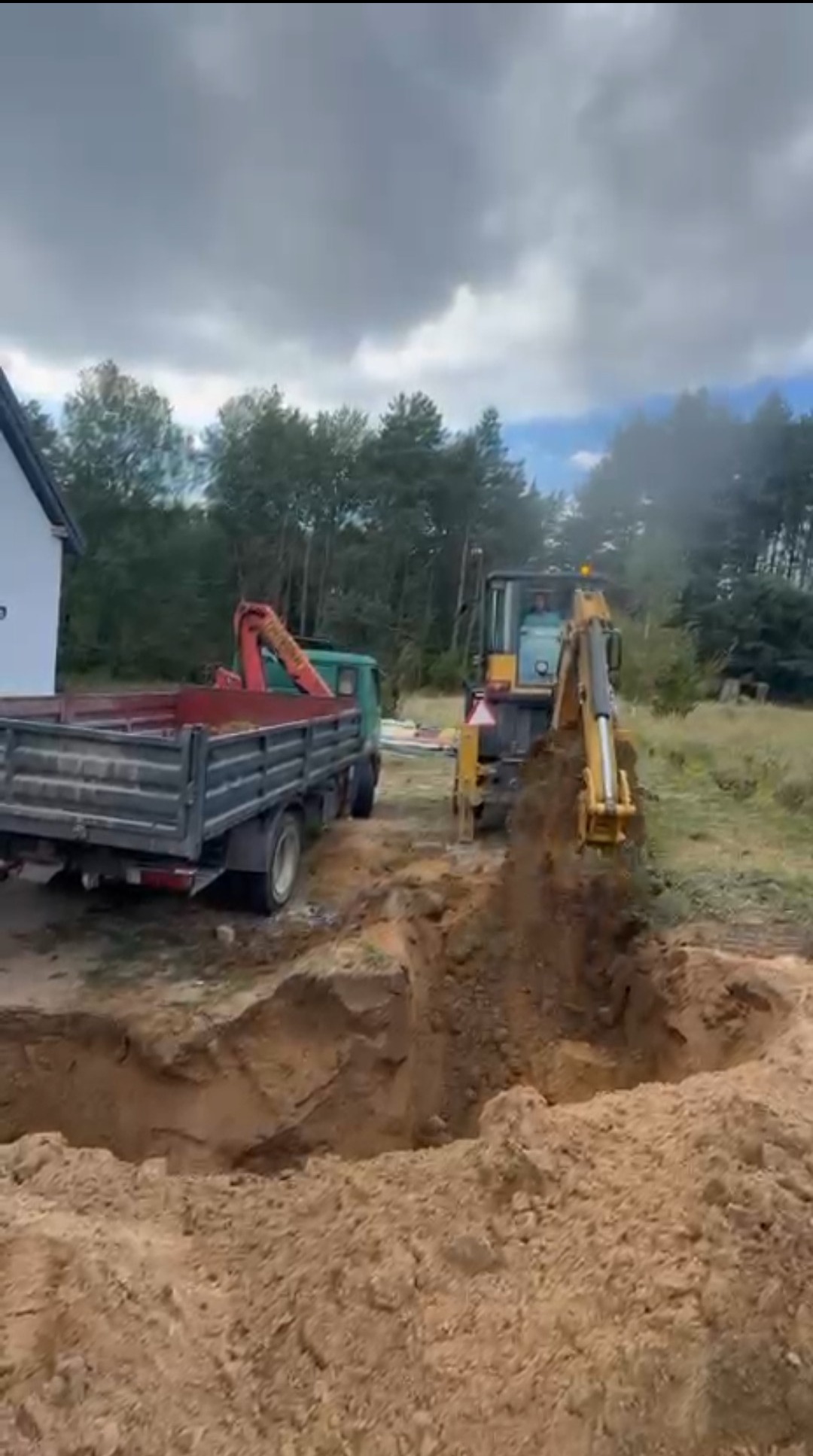 Koparka wykopuje dół w ziemi obok zaparkowanej ciężarówki z dźwigiem, prawdopodobnie pod fundamenty lub instalację. Widoczne drzewa i fragment budynku w tle.