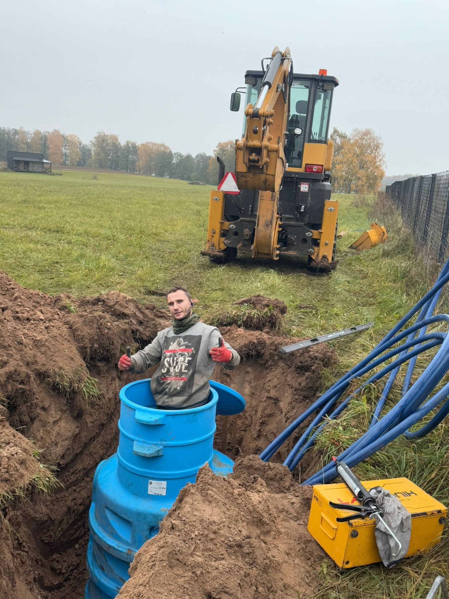Montaż niebieskiego zbiornika w wykopie, z pracownikiem dającym kciuki w górę, koparką w tle i niebieskimi rurami obok. Prace ziemne i instalacyjne w toku.