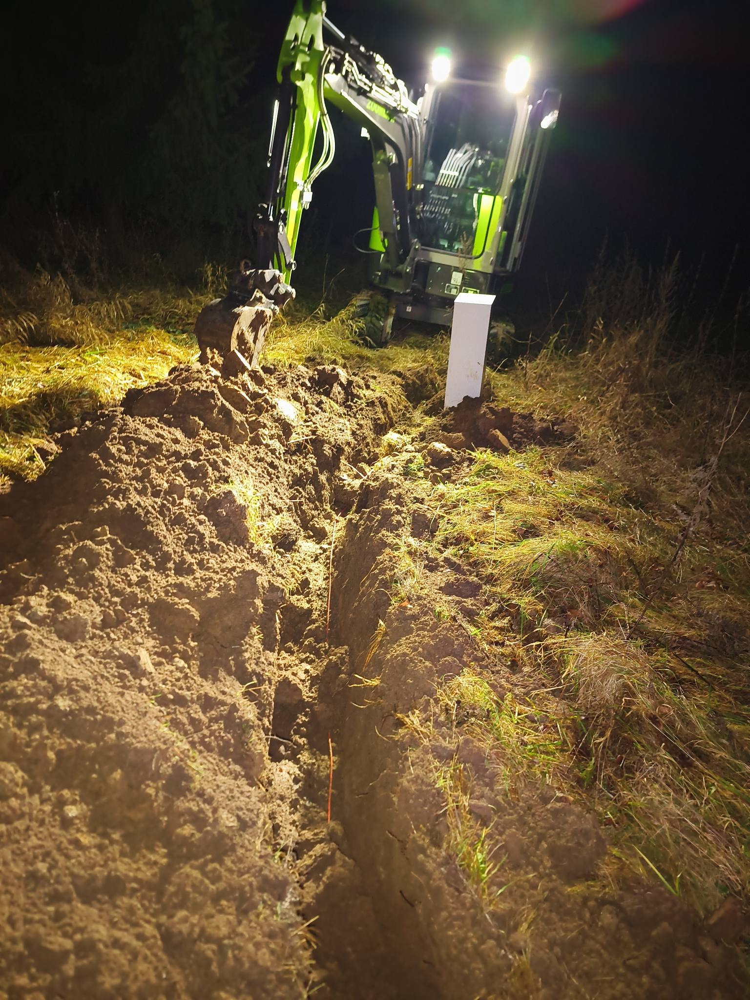 Nocne prace ziemne: zielona koparka wykopuje wąski rów w ziemi, oświetlona reflektorami. W tle widoczna roślinność i biały słupek.