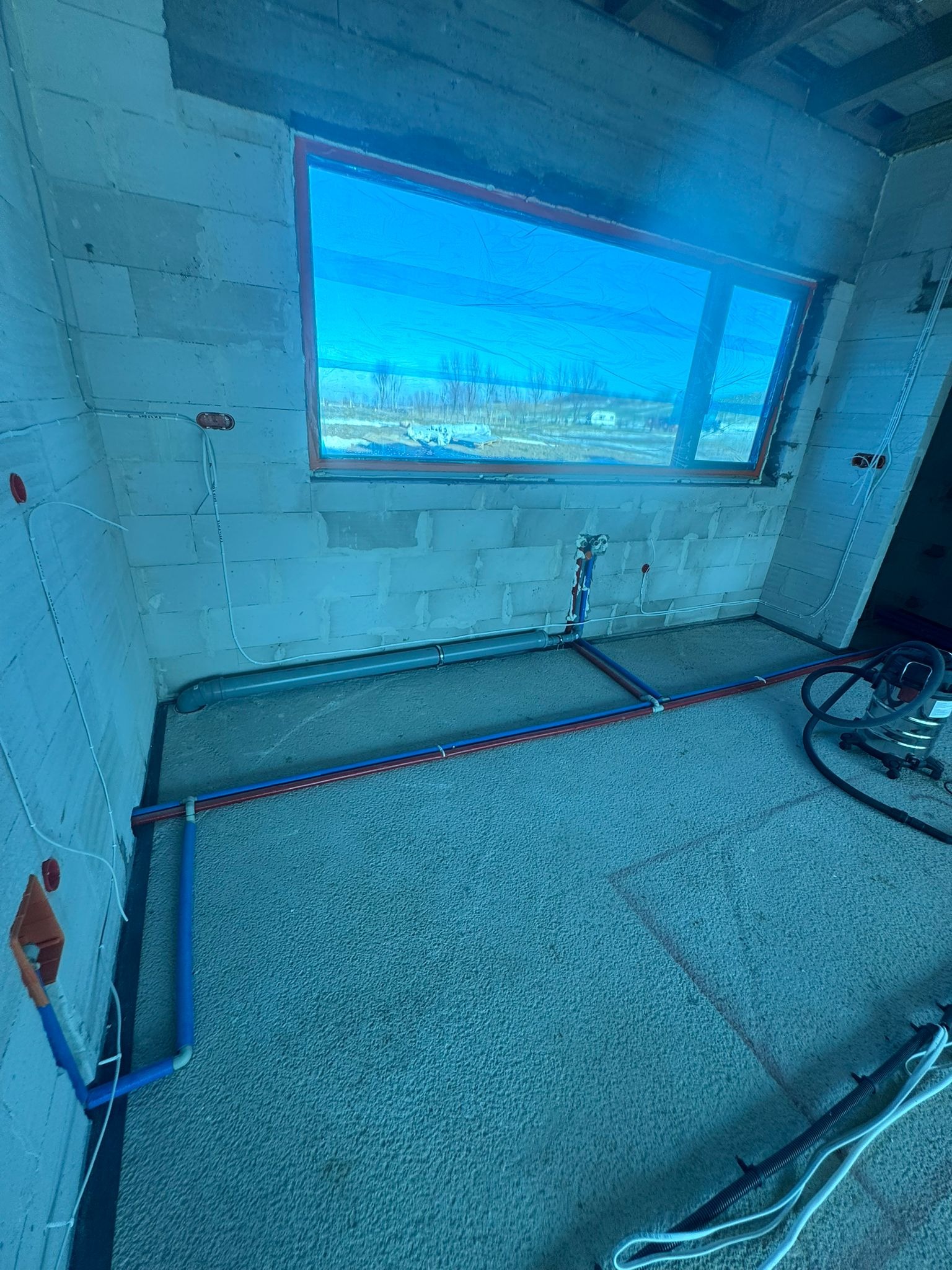 Instalacja wodna podłogowa w surowym wnętrzu z oknem. Rury w kolorze niebieskim i czerwonym ułożone na szarym podłożu. Widoczne przewody elektryczne na ścianach.
