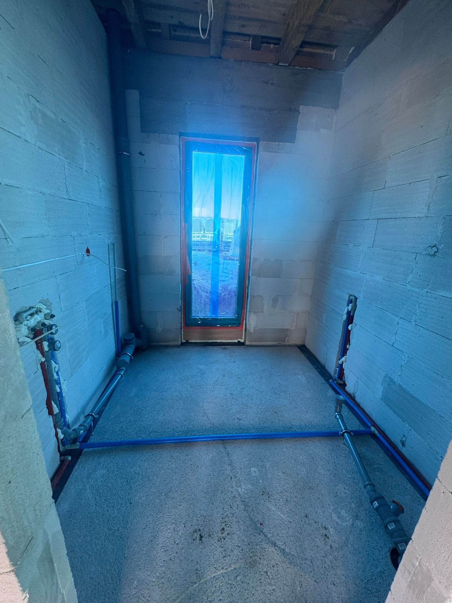 Wnętrze w trakcie instalacji hydraulicznej: widoczne rury PEX w podłodze i przy ścianach z bloczków, okno zabezpieczone folią. Etap prac przed wylewką.