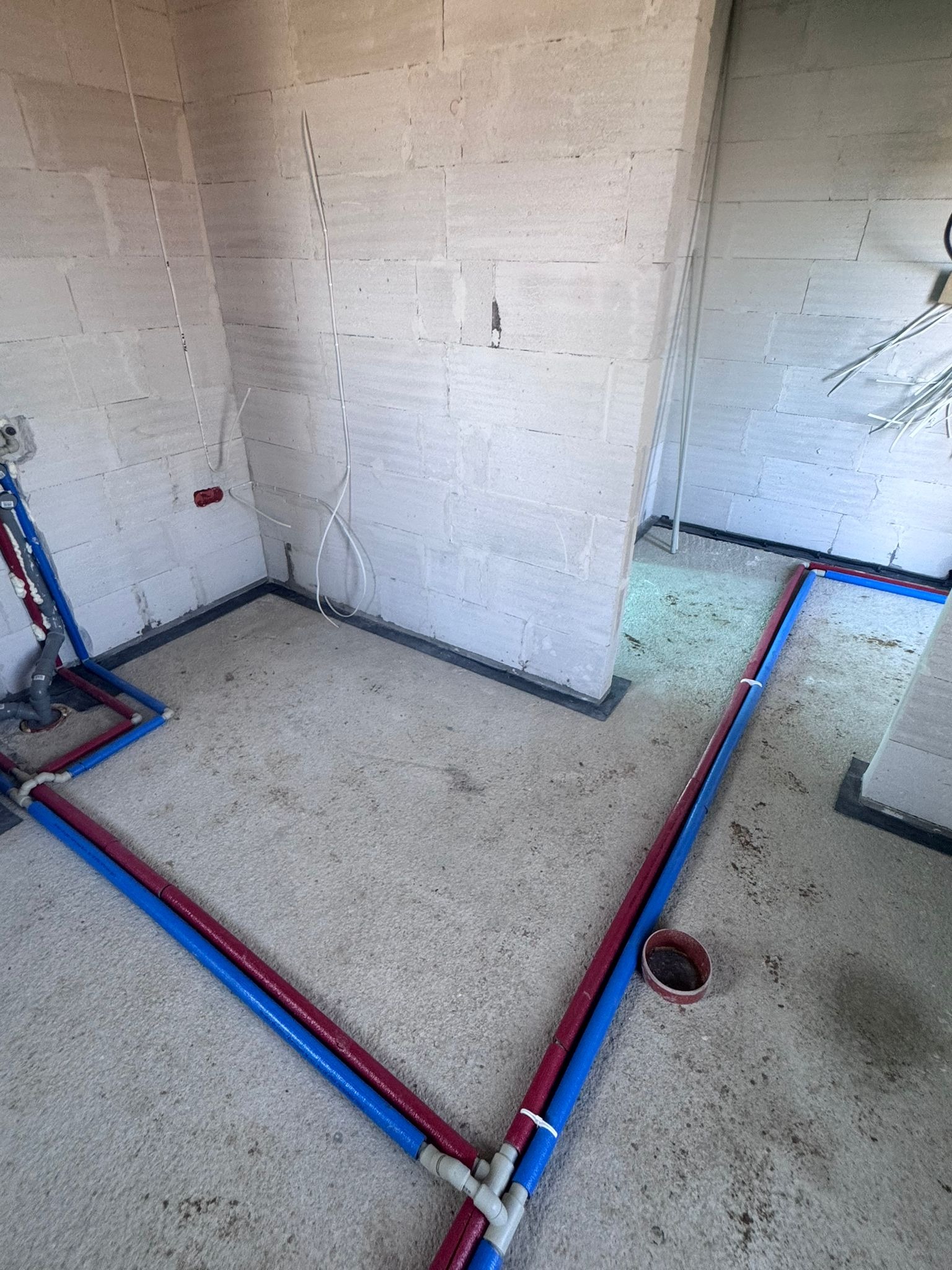 Instalacja wodna w surowym wnętrzu: czerwone i niebieskie rury prowadzone po podłodze wzdłuż ścian z betonu komórkowego, widoczne złącza i fragment instalacji przy ścianie.