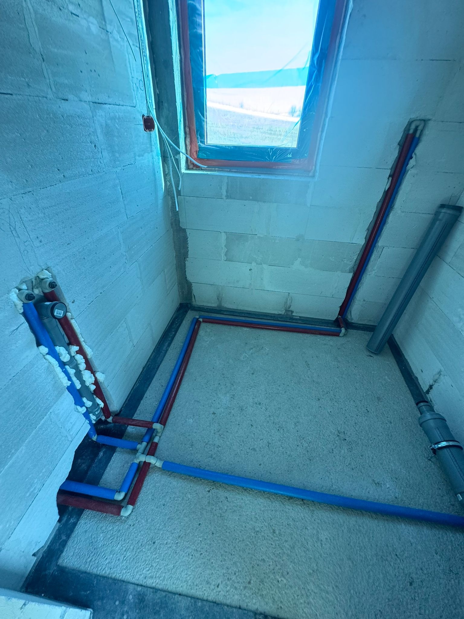Wnętrze w trakcie instalacji hydraulicznej: rury (czerwone, niebieskie) prowadzone wzdłuż ścian i podłogi, okno z widokiem na zewnątrz, ściany z białych bloczków.