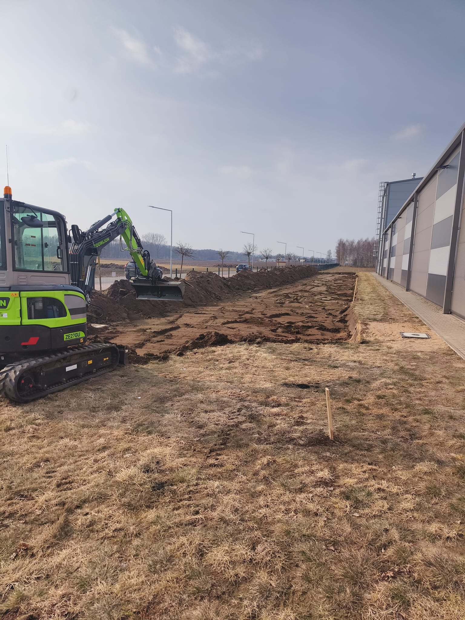 Zielona koparka Zoomlion ZE27GU w trakcie wykopu długiego rowu w ziemi, obok budynku z szarej blachy trapezowej. Widoczne drzewa w tle.