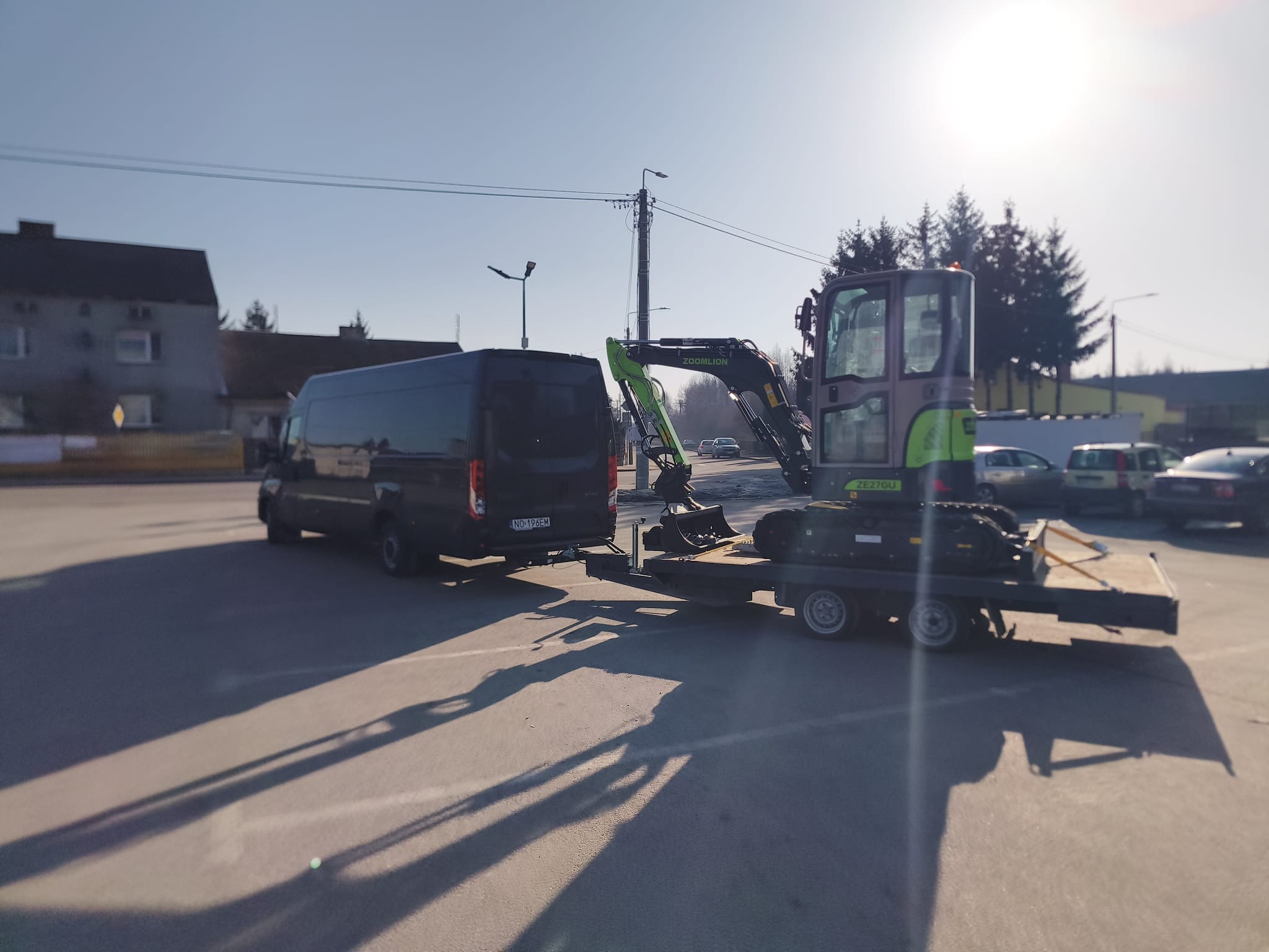 Czarny bus z przyczepą przewożącą minikoparkę Zoomlion ZE27GU na tle miejskiego krajobrazu w słoneczny dzień. Długie cienie pojazdów na asfalcie.