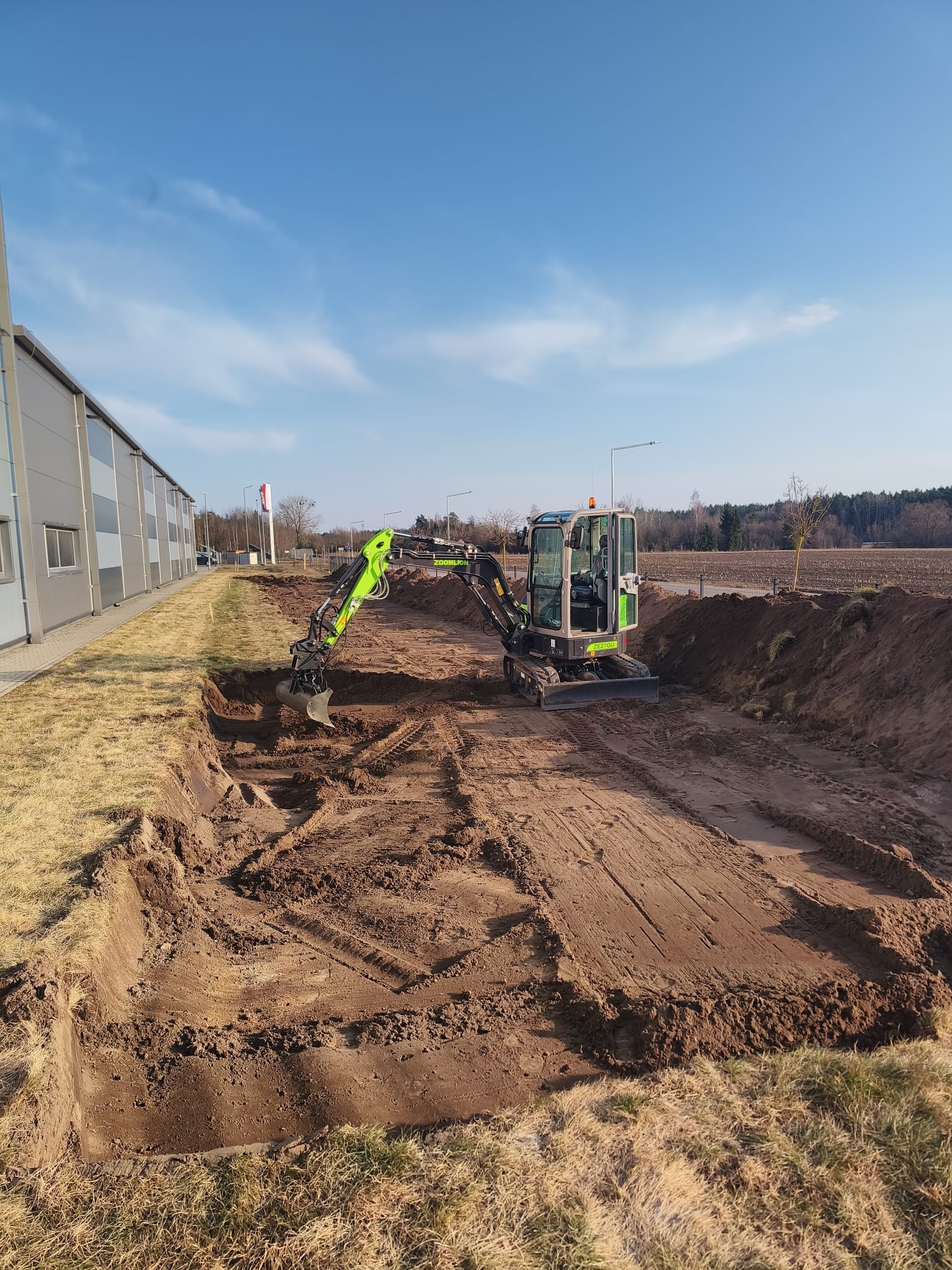 Mini koparka Zoomlion w trakcie wykopu prostokątnego dołu w ziemi, obok budynku przemysłowego, w słoneczny dzień. Widoczne ślady opon i świeżo wykopana ziemia.