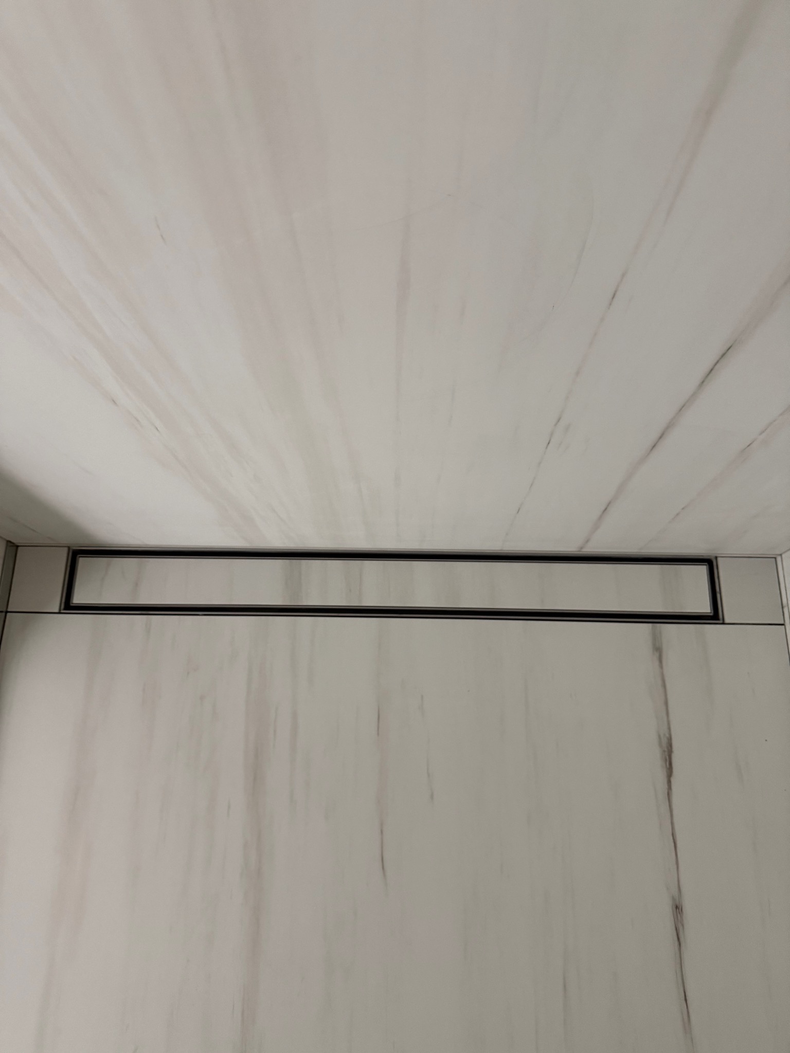 Białe płytki imitujące marmur z czarnym, liniowym odpływem w podłodze kabiny prysznicowej. Minimalistyczny design, nowoczesne wykończenie łazienki.