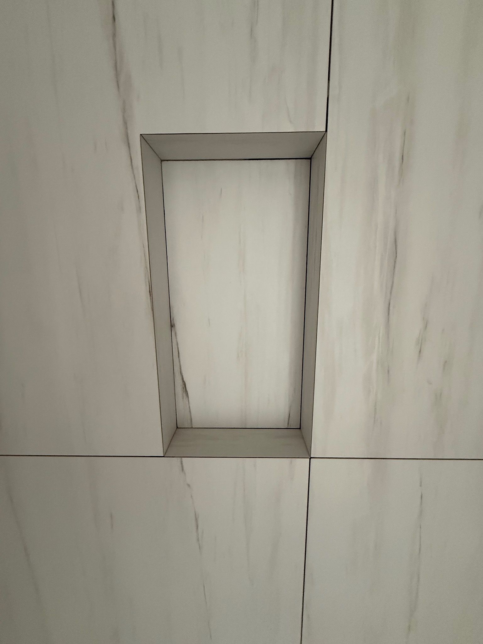 Wnęka w ścianie wyłożonej białymi płytkami imitującymi marmur. Wnęka ma prostokątny kształt z delikatnie trapezowymi bokami, obramowana szarą listwą.