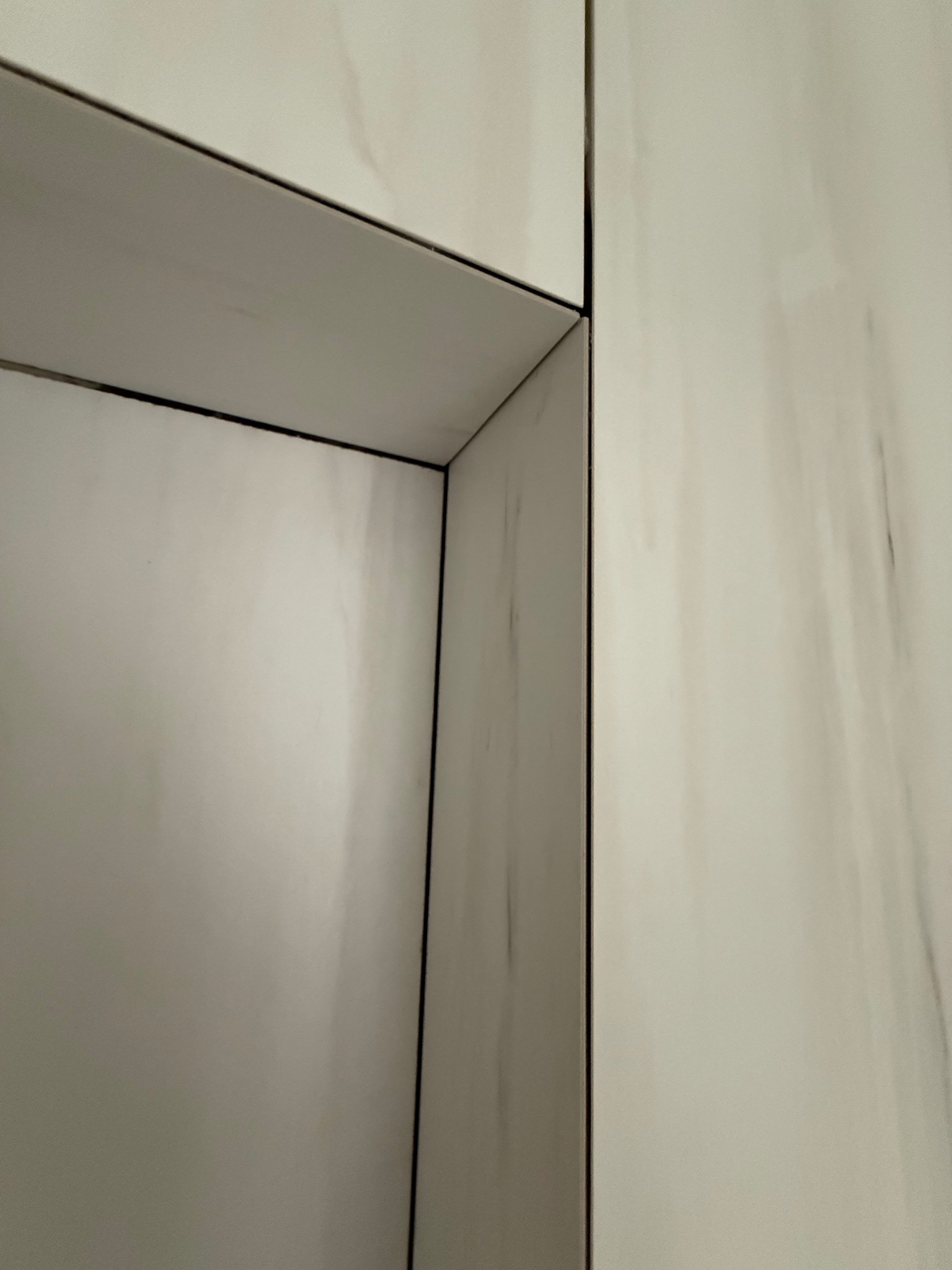 Narożnik wykończony białymi płytkami imitującymi marmur, z ciemną fugą, tworzący wnękę. Precyzyjne ułożenie płytek, widoczne detale wykończenia.