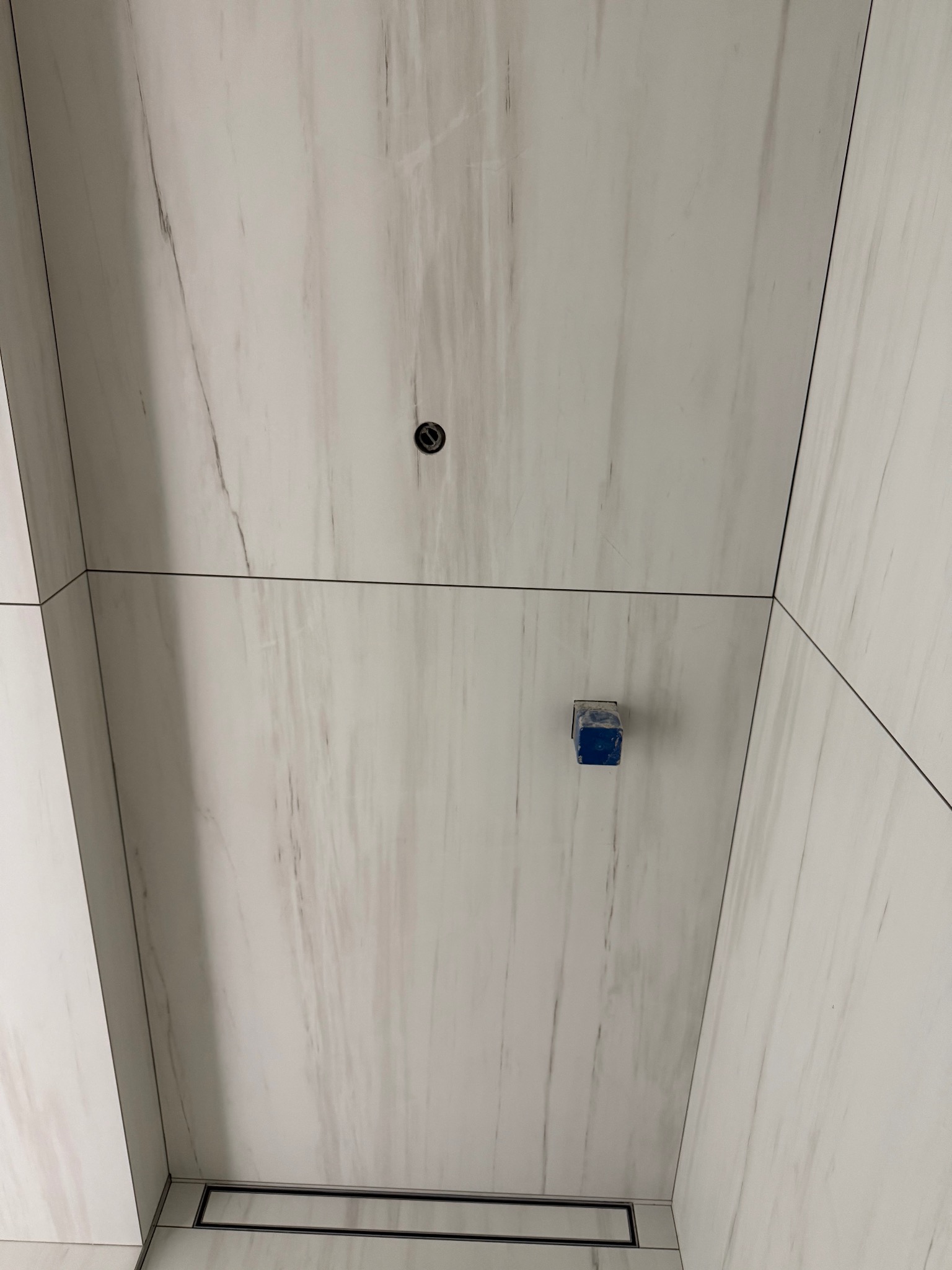 Minimalistyczna łazienka z białymi płytkami imitującymi marmur, czarnym odpływem liniowym i punktowym oświetleniem. Widok z dołu, podkreślający geometrię i detale wykończenia.