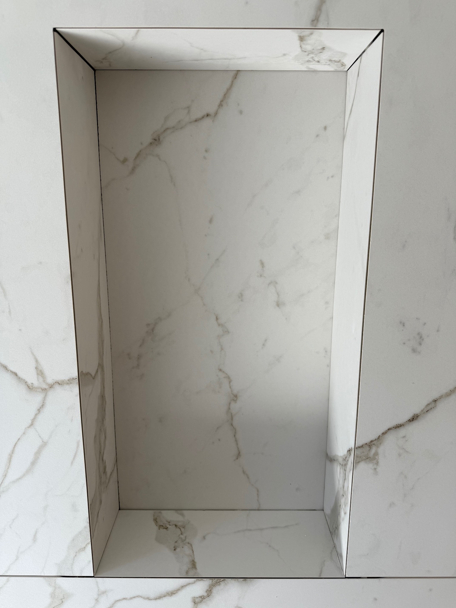 Narożna wnęka w łazience wyłożona białymi płytkami imitującymi marmur, z precyzyjnie dociętymi krawędziami i fugami. Minimalistyczny design, nowoczesne wykończenie.