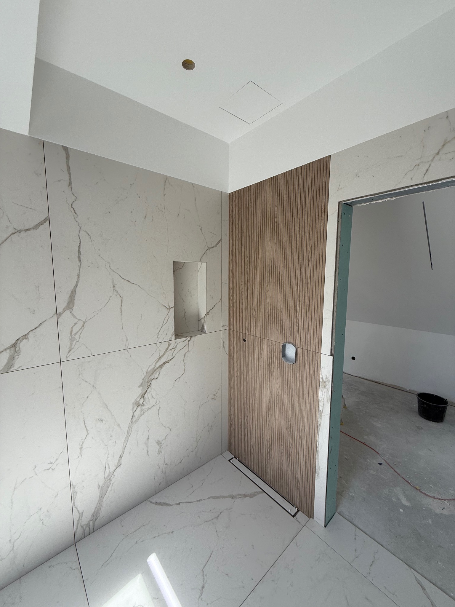 Wykończona łazienka z marmurowymi płytkami, drewnianą ścianą i odpływem liniowym. Widoczny fragment sufitu z oświetleniem i otwór drzwiowy do innego pomieszczenia.