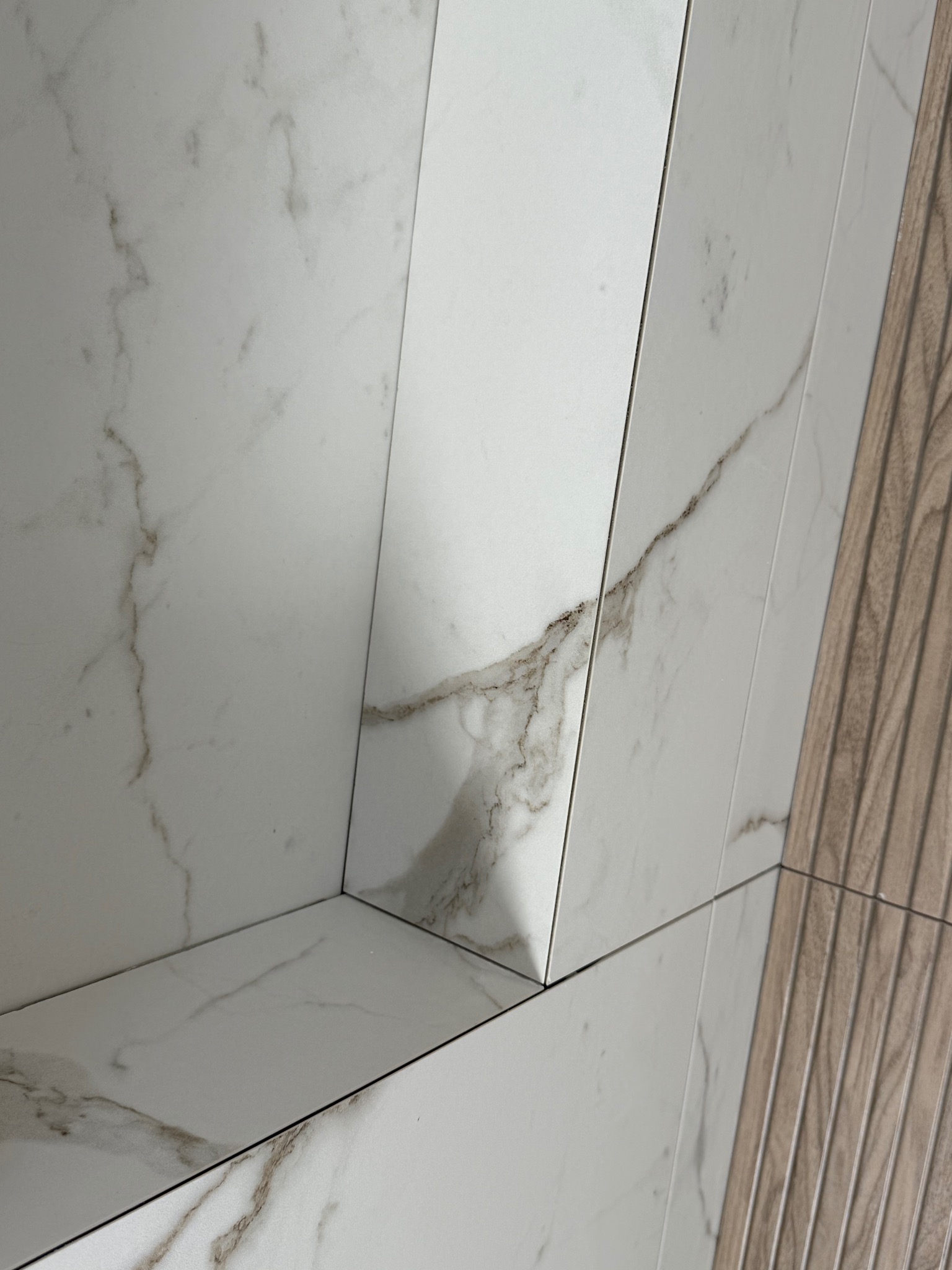 Wykończona wnęka z białych płytek imitujących marmur, sąsiadująca z pionowymi panelami ściennymi o strukturze drewna. Detal wykończenia łazienki.