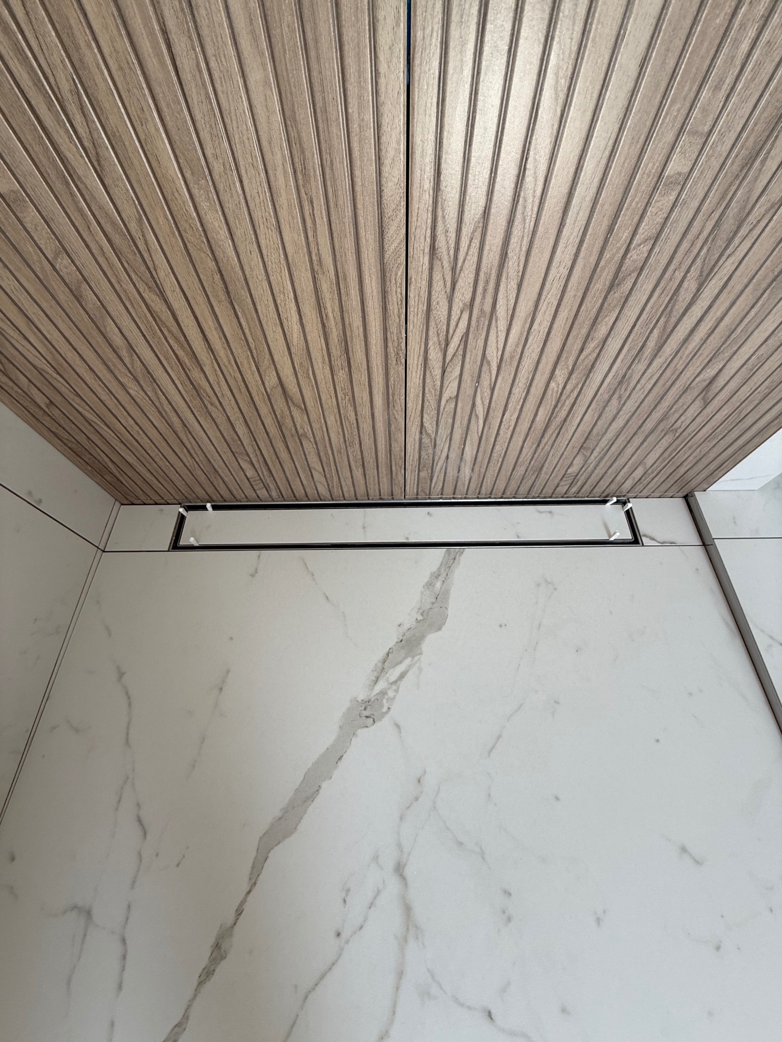 Nowoczesna łazienka z drewnianym sufitem lamelowym i odpływem liniowym w posadzce z białego marmuru z szarymi żyłami.