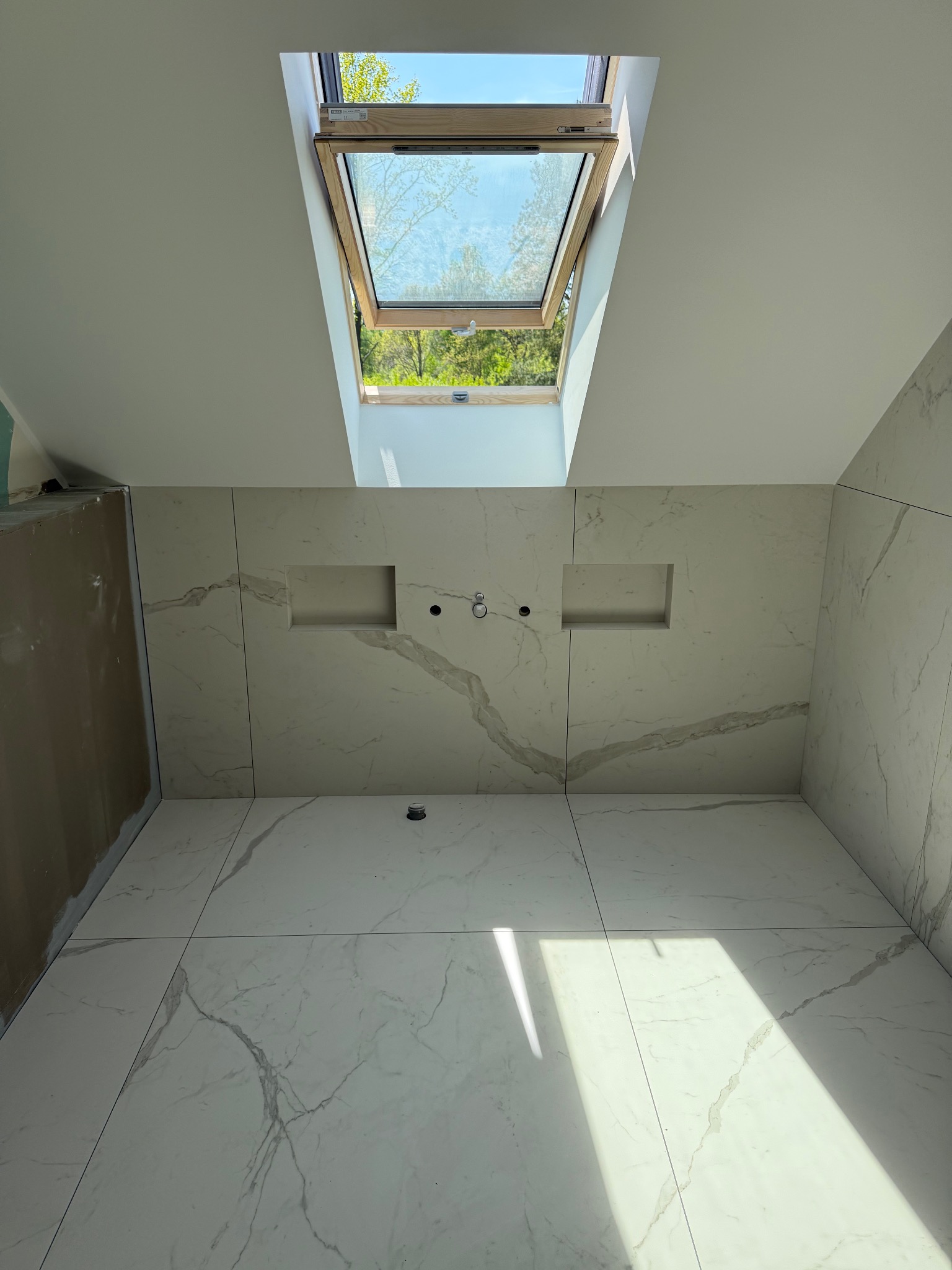 Wykończona łazienka z białymi płytkami imitującymi marmur, wnękami w ścianie i oknem dachowym wpuszczającym naturalne światło. Widok z dołu, podkreślający przestronność.
