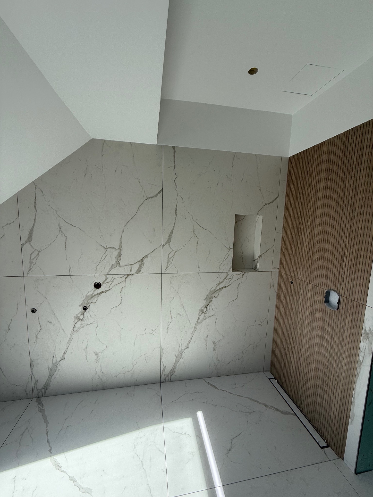 Wykończona łazienka z marmurowymi płytkami i drewnianą ścianą z ukrytymi otworami na armaturę. Widoczny odpływ liniowy i wnęka w ścianie.
