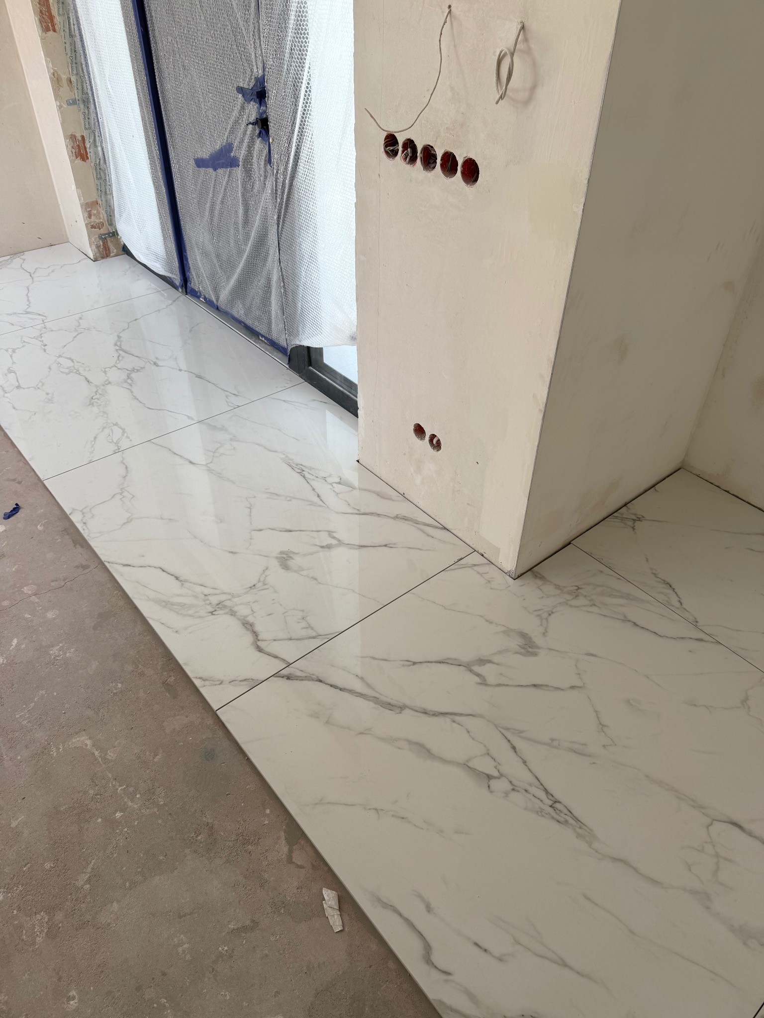 Ułożone białe płytki imitujące marmur na podłodze w trakcie remontu. Widoczne przygotowane otwory na gniazdka elektryczne i zabezpieczone folią drzwi balkonowe.