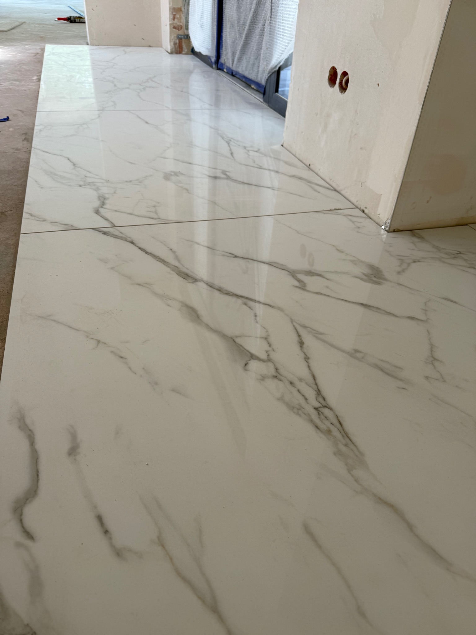 Błyszcząca, biała podłoga z płytek imitujących marmur, świeżo ułożona w pomieszczeniu w trakcie wykańczania. Widoczne gniazdka elektryczne w ścianie.