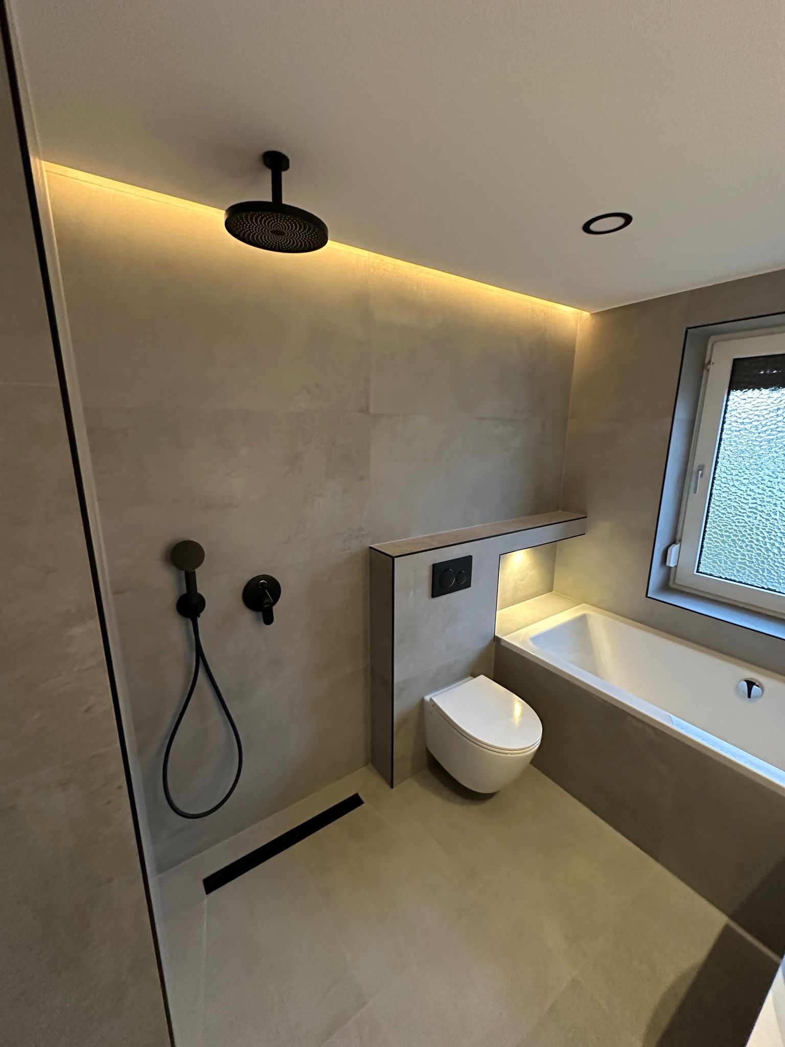 Nowoczesna łazienka w odcieniach beżu z czarnymi akcentami: deszczownica, bateria, odpływ liniowy. Minimalistyczny design, podświetlenie LED, wanna i podwieszana toaleta.