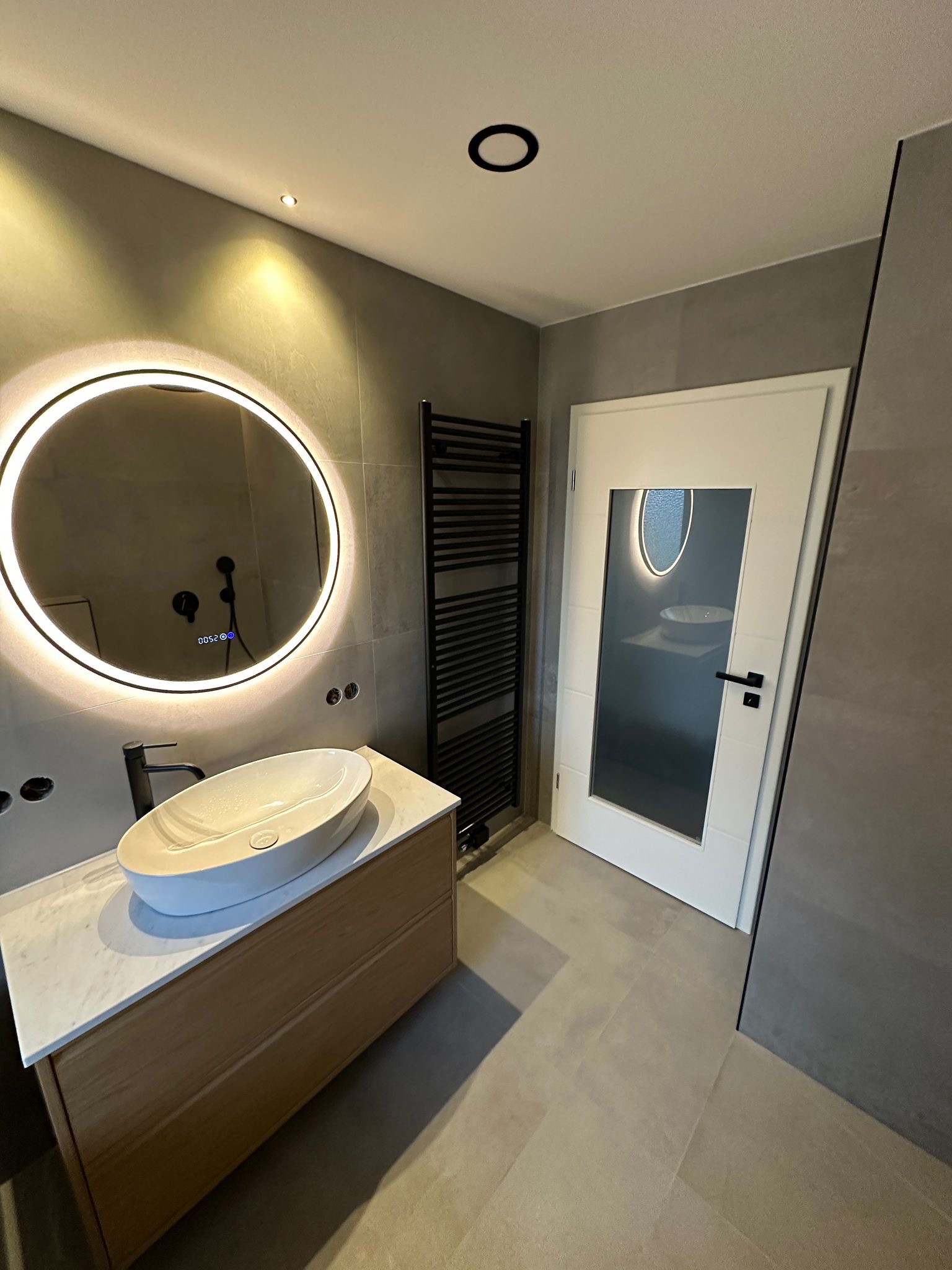 Nowoczesna łazienka z owalną umywalką, podświetlanym lustrem i grzejnikiem drabinkowym. Szare płytki i białe drzwi z owalnym przeszkleniem tworzą spójną całość.