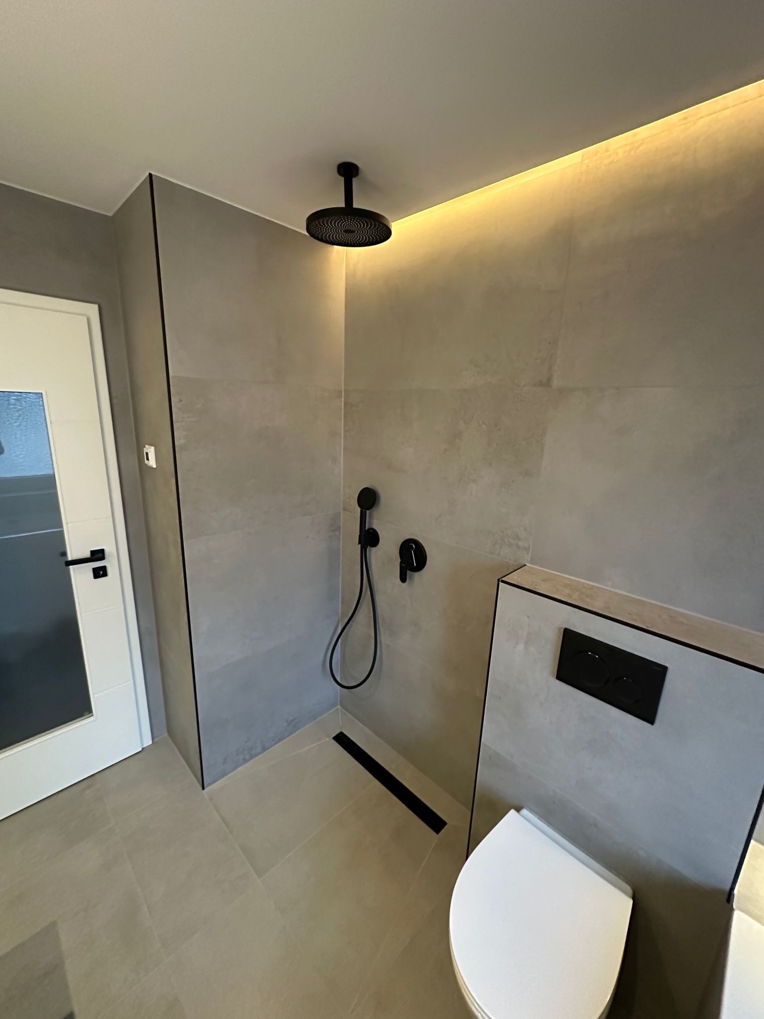 Nowoczesna łazienka z szarymi płytkami, czarną armaturą i podświetleniem LED. Minimalistyczny design z kabiną prysznicową typu walk-in i podwieszaną toaletą.