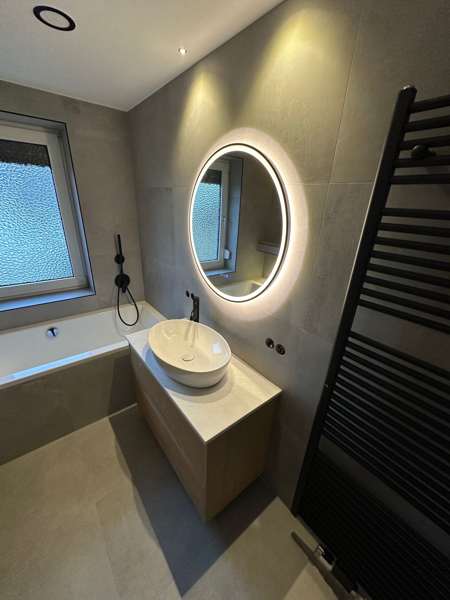 Nowoczesna łazienka z owalną umywalką, podświetlanym lustrem i grzejnikiem drabinkowym. Szare płytki, minimalistyczny design, ciepłe oświetlenie punktowe.