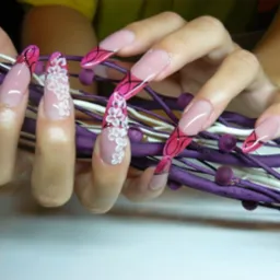 Dłoń z długimi paznokciami, zdobionymi różowym lakierem, czarnymi liniami i trójwymiarowymi białymi kwiatkami, na tle fioletowych gałązek.