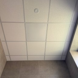 MTG USŁUGI BUDOWLANE - Kasetonowy sufit podwieszany z widocznym czujnikiem dymu i płytą rewizyjną, połączony z wykończoną płytkami ścianą. Ujęcie z dołu, podkreślające regularność wzoru.