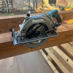 MTG USŁUGI BUDOWLANE - Elektryczna piła tarczowa Makita na brązowych belkach drewnianych, w tle budynek z drewnem opałowym. Narzędzie do cięcia drewna w budownictwie.