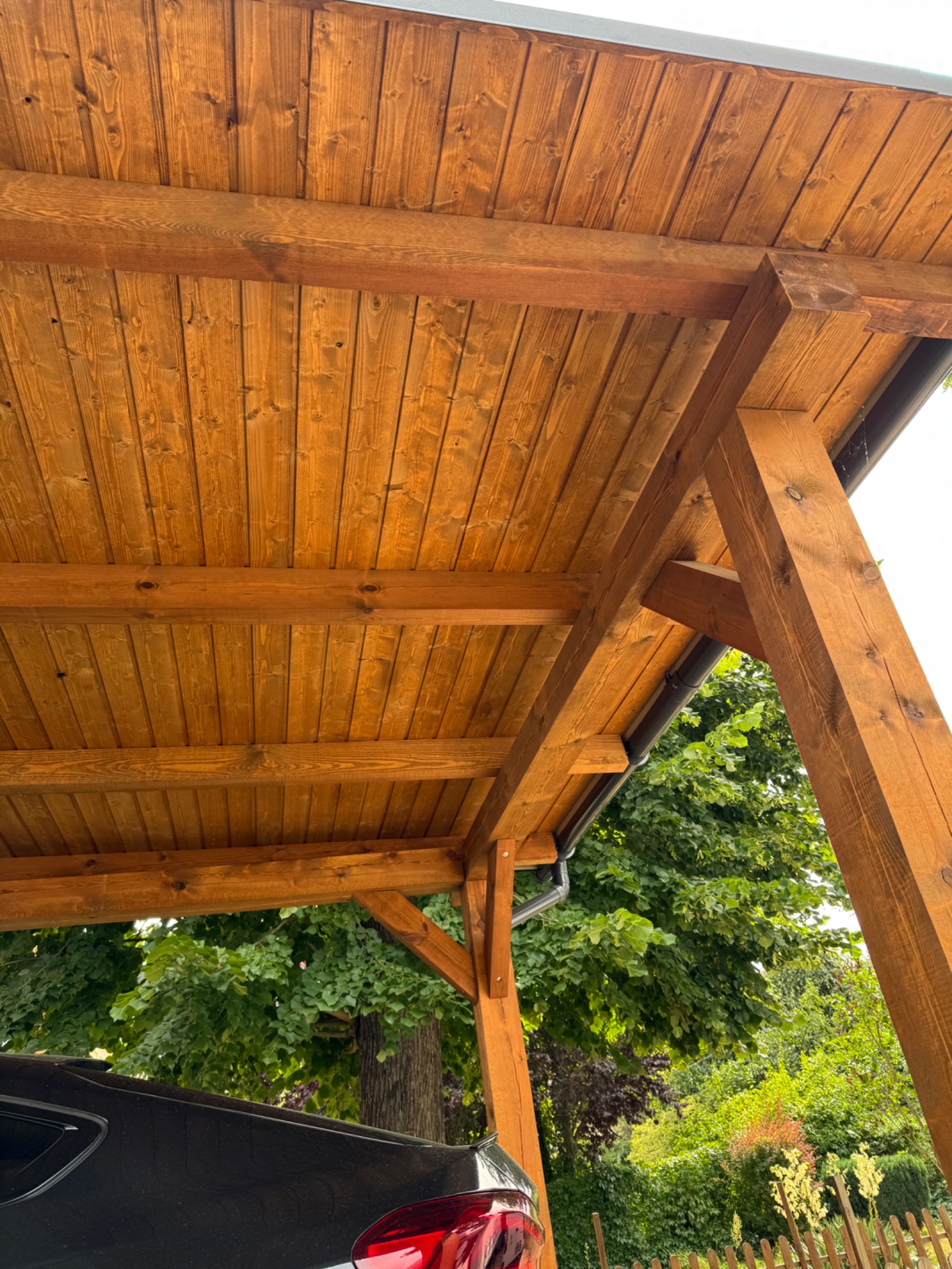 Drewniana wiata garażowa z widoczną konstrukcją dachu i fragmentem samochodu. Naturalne oświetlenie, widok na zieleń w tle. Solidne wykonanie, detale ciesielskie.