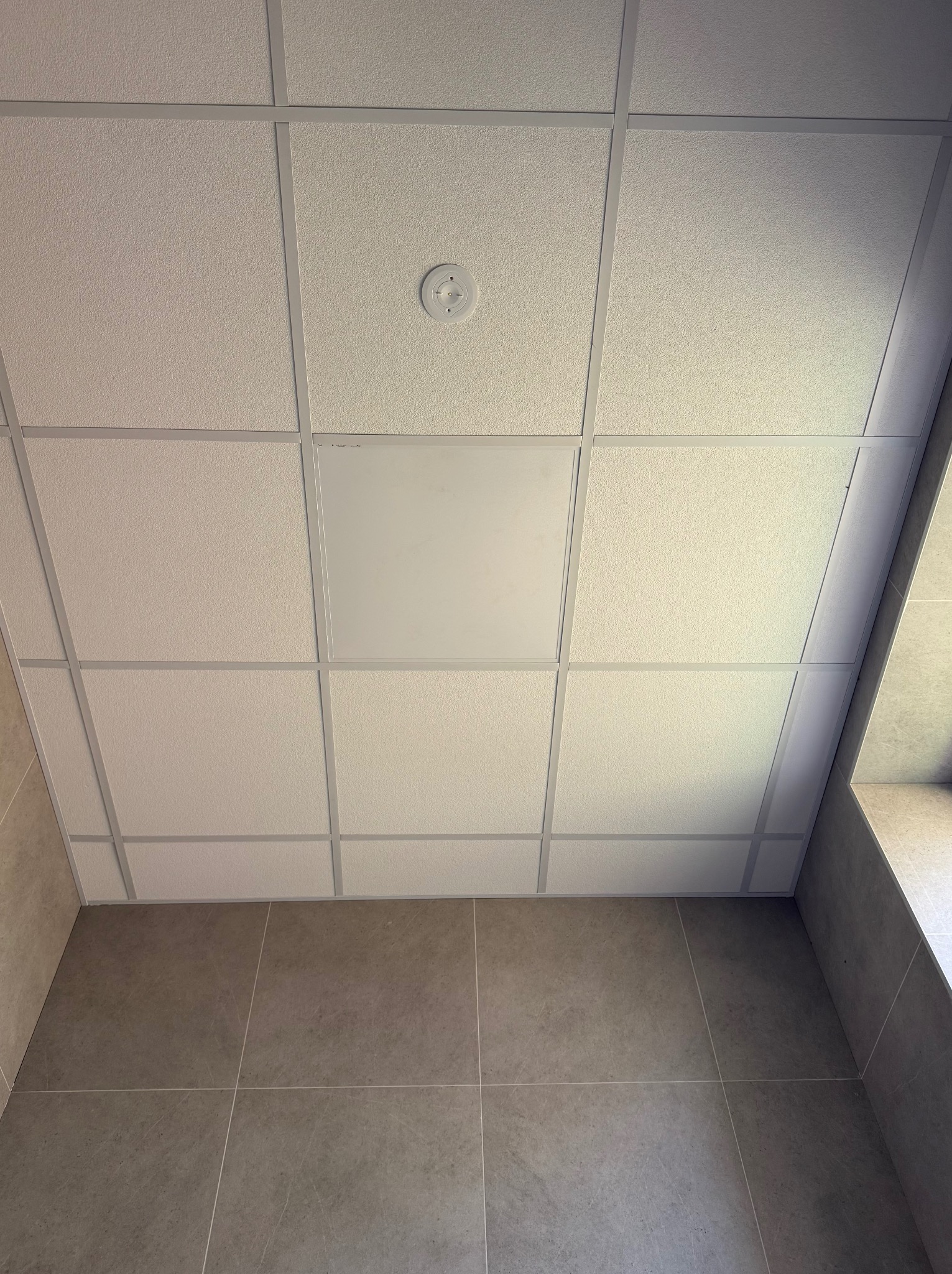 Kasetonowy sufit podwieszany z widocznym czujnikiem dymu i płytą rewizyjną, połączony z wykończoną płytkami ścianą. Ujęcie z dołu, podkreślające regularność wzoru.