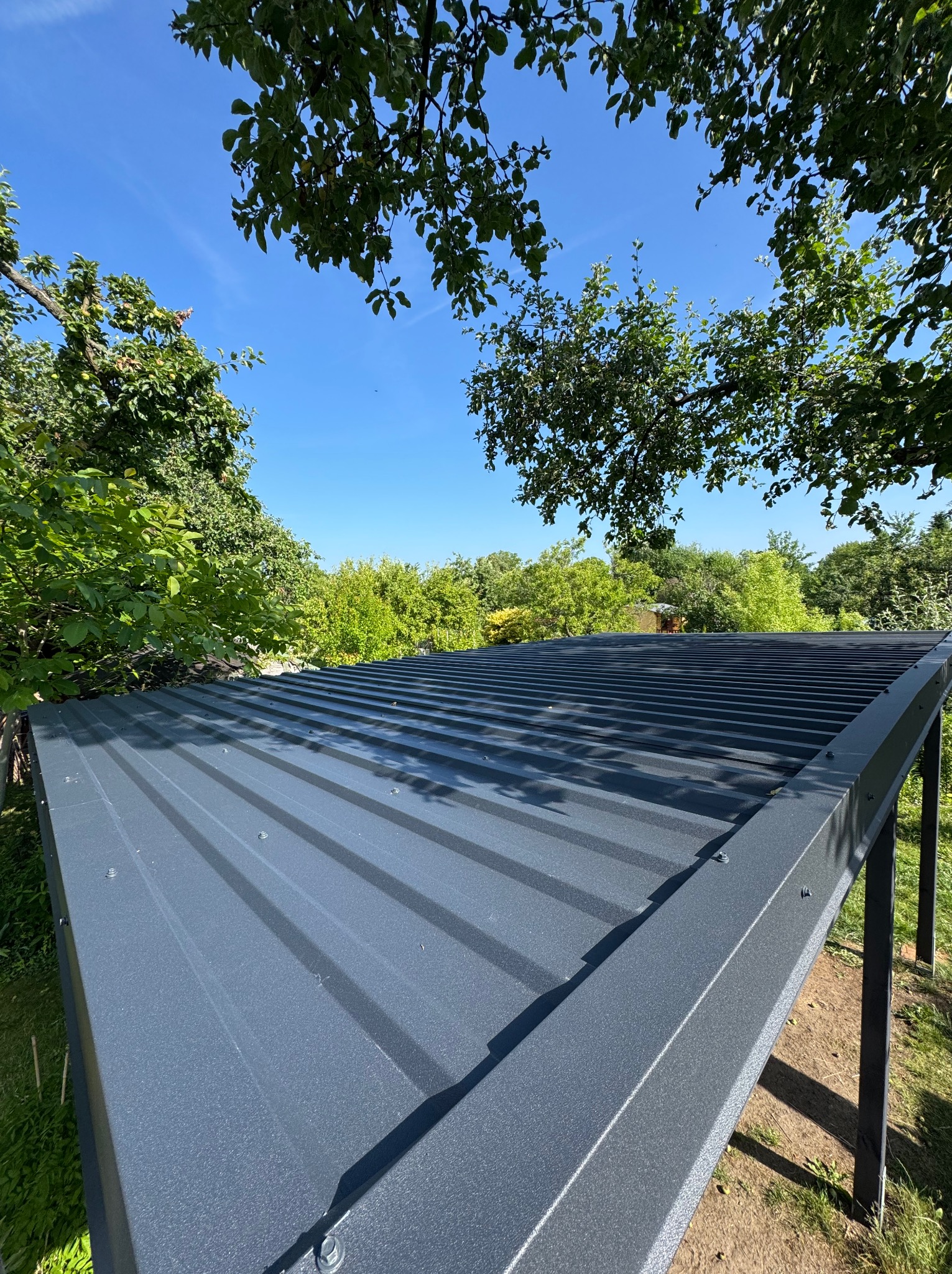 Metalowa konstrukcja z zadaszeniem w ogrodzie, widok z dołu na tle zieleni i błękitnego nieba. Elementy złączne widoczne na krawędziach dachu.