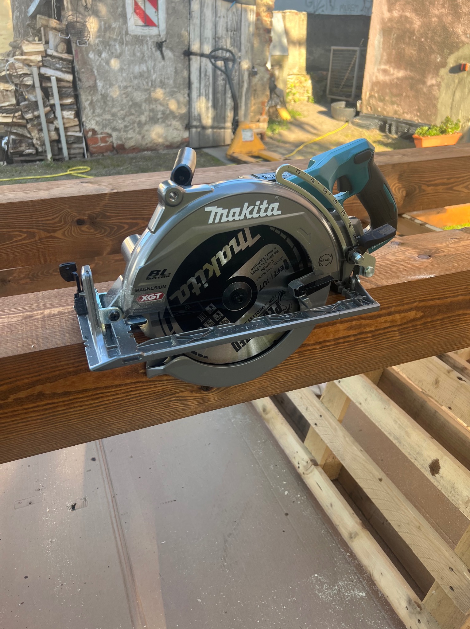 Elektryczna piła tarczowa Makita na brązowych belkach drewnianych, w tle budynek z drewnem opałowym. Narzędzie do cięcia drewna w budownictwie.