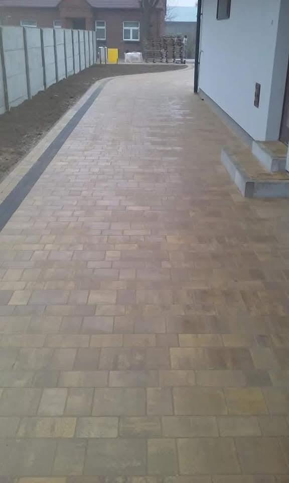 Ułożona, beżowa kostka brukowa tworząca ścieżkę obok domu z szarym obramowaniem. W tle widoczne ogrodzenie z betonowych płyt i palety z kostką.