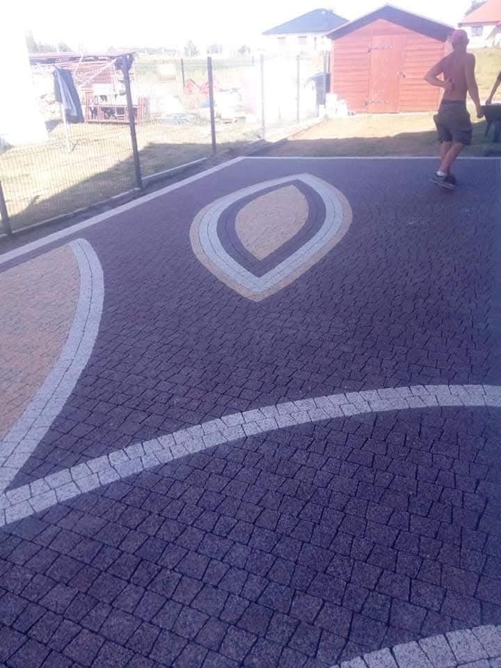 Ułożona kostka brukowa w ciemnych odcieniach z dekoracyjnymi wzorami w kształcie łezek i łuków z jasnej kostki. W tle widoczny fragment ogrodzenia i budynek.