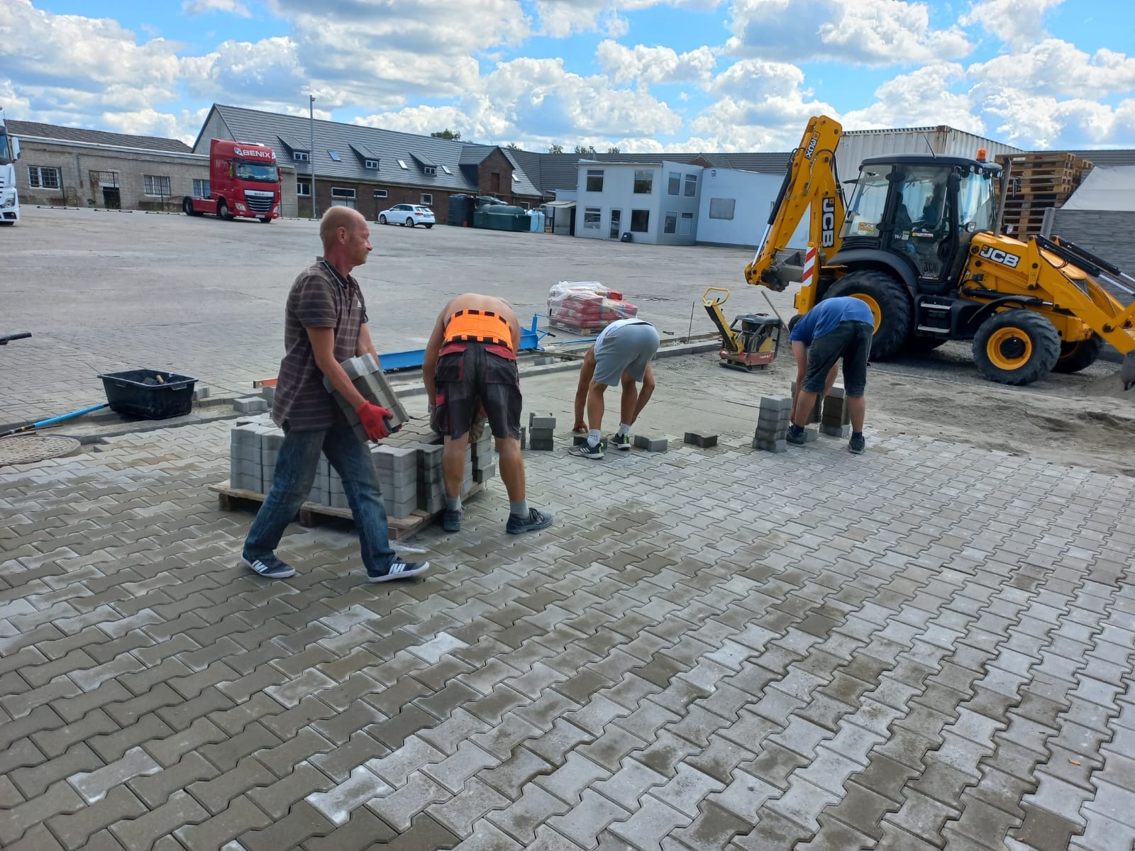 Układanie kostki brukowej na placu: czterech mężczyzn pracuje, jeden transportuje kostkę, pozostali układają. W tle koparka JCB i budynki.
