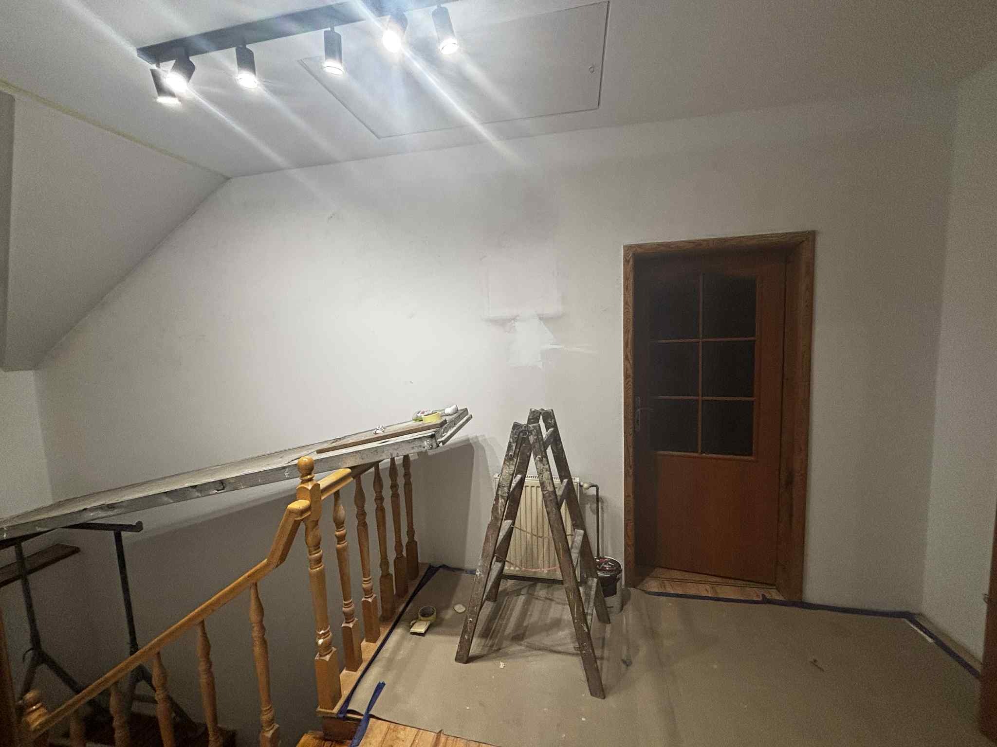 Wnętrze w trakcie malowania: drabina, wałek i zabezpieczona podłoga. Ściana częściowo pomalowana na biało, widoczne ślady po szpachlowaniu. Oświetlenie sufitowe.