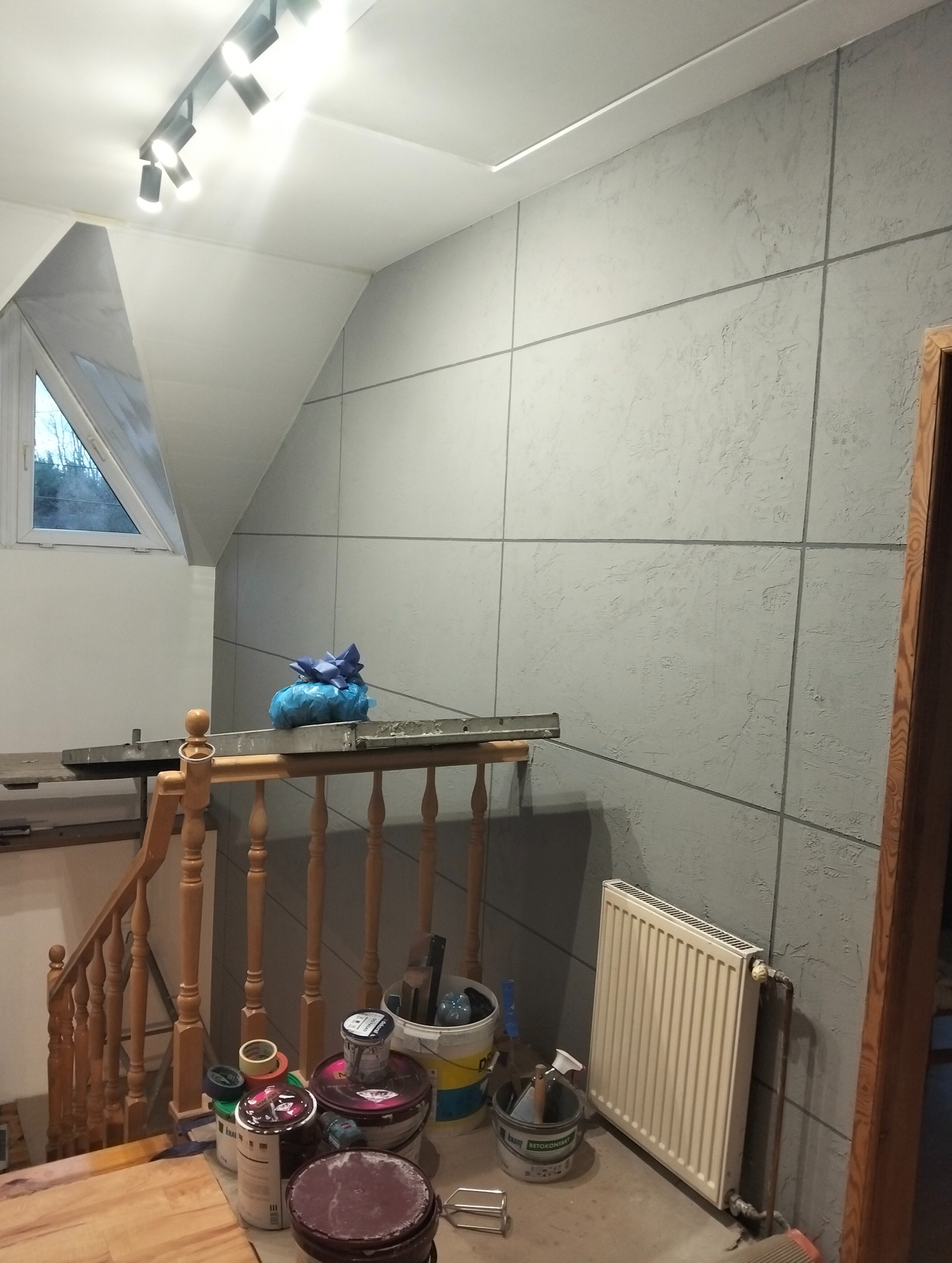 Ściana z szarą, strukturalną okładziną w kwadraty, widoczna klatka schodowa z drewnianą balustradą, puszki z farbą i grzejnik. Ukończone wykończenie wnętrza.