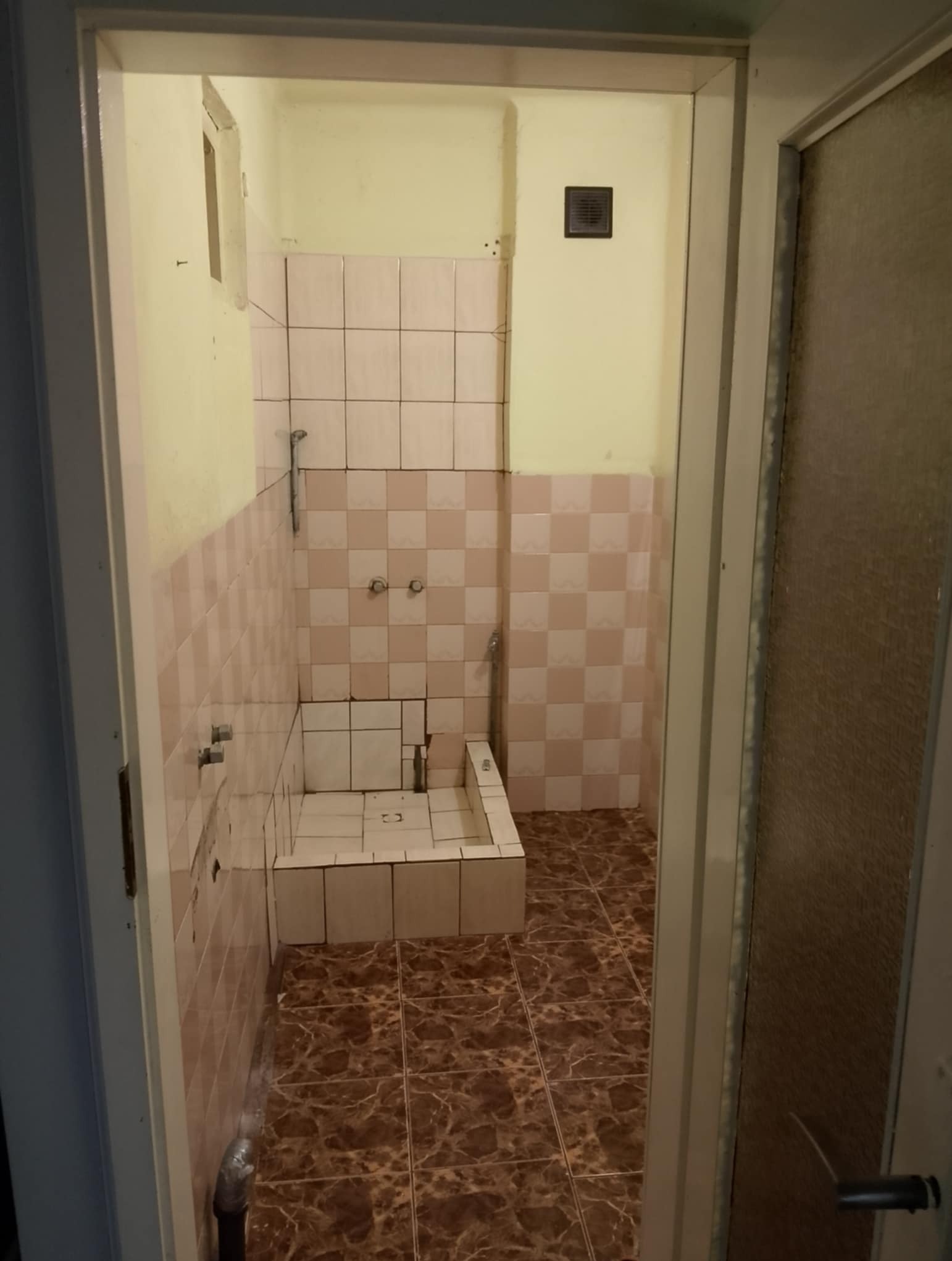 Wnętrze łazienki z brudnymi, różowo-białymi płytkami i brązową podłogą, widziane zza otwartych drzwi. Widoczny fragment wanny i ściany z armaturą.