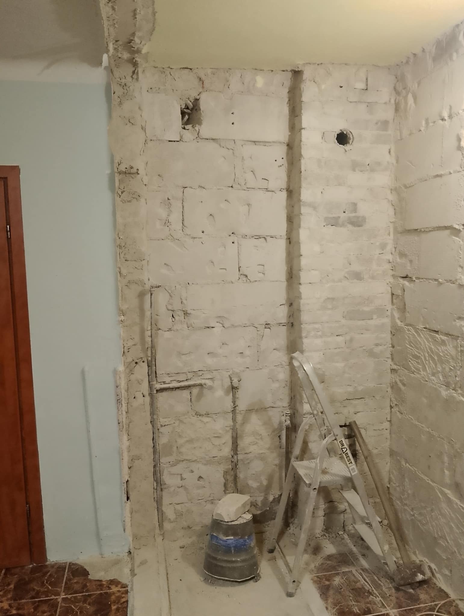 Ściana z surowej cegły w trakcie prac przygotowawczych do wykończenia, widoczna drabina i wiadro z zaprawą. Fragment podłogi z terakoty.