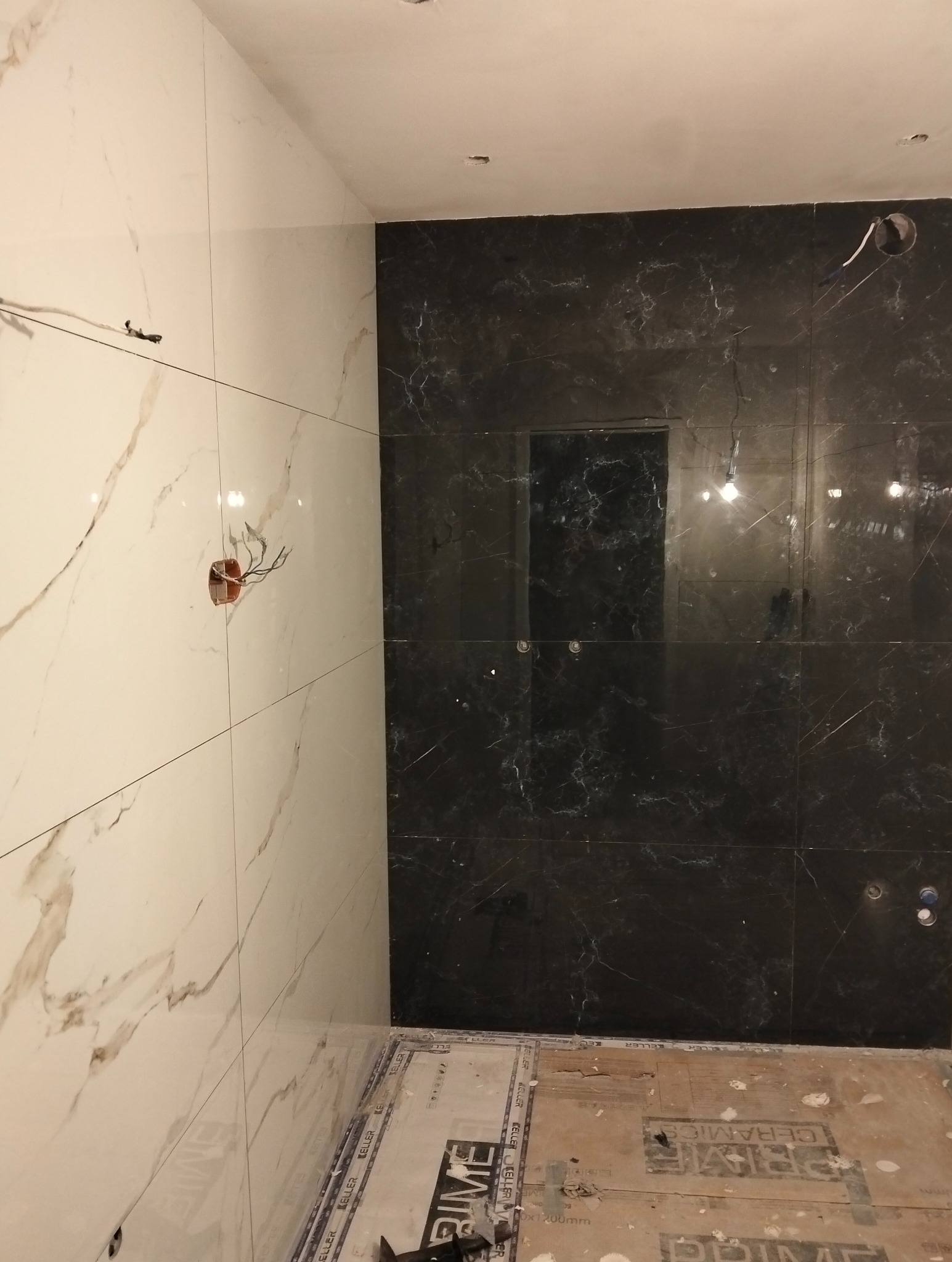 Częściowo wykończona łazienka z białymi i czarnymi płytkami imitującymi marmur. Widoczne otwory na instalacje elektryczne i sanitarne. Podłoga zabezpieczona folią.