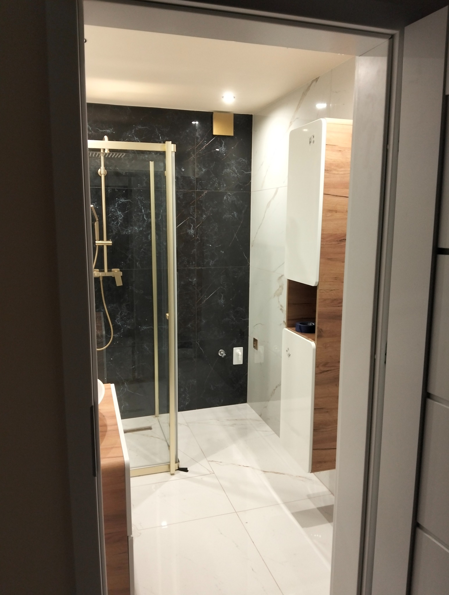Nowoczesna łazienka z kabiną prysznicową w złotej ramie, czarnymi marmurowymi płytkami i białymi płytkami z szarymi żyłami. Widok zza drzwi.