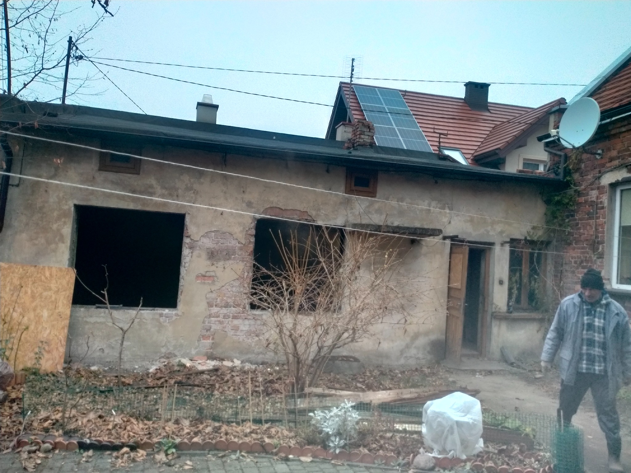 Niski budynek z odsłoniętą cegłą i pustymi otworami okiennymi, widoczny mężczyzna w tle, sugerujący prace remontowe lub przygotowawcze do nich.