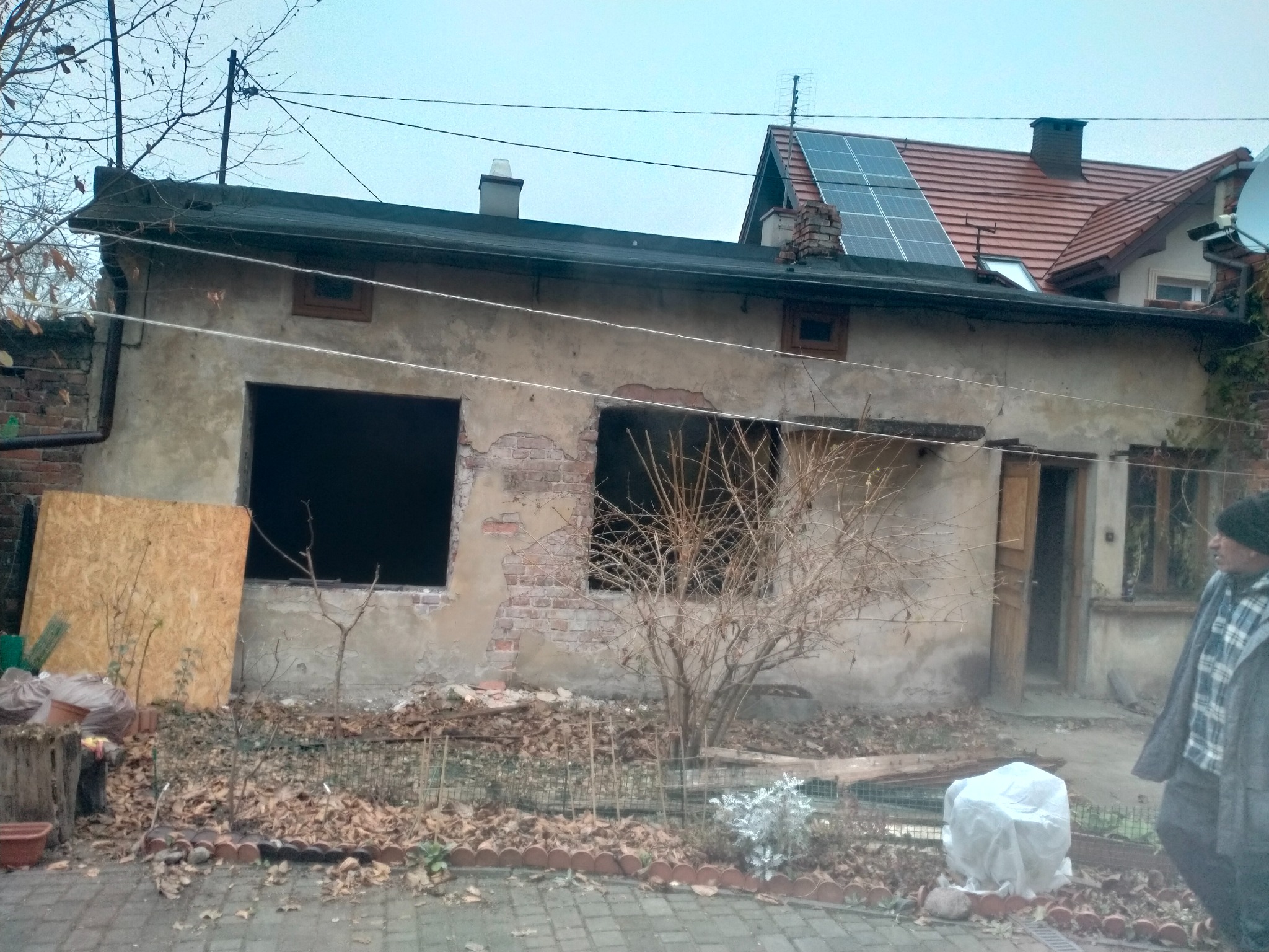 Dom przed remontem z odsłoniętą cegłą, niezabezpieczonymi oknami i panelami słonecznymi na dachu. Na pierwszym planie liście i osoba w kurtce.