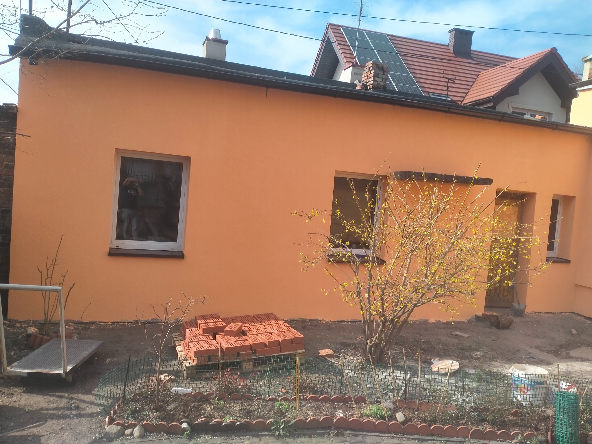 Otynkowany budynek w odcieniach pomarańczy z widocznymi oknami i stertą cegieł. W tle dom z panelami słonecznymi. Widoczny fragment ogrodu.