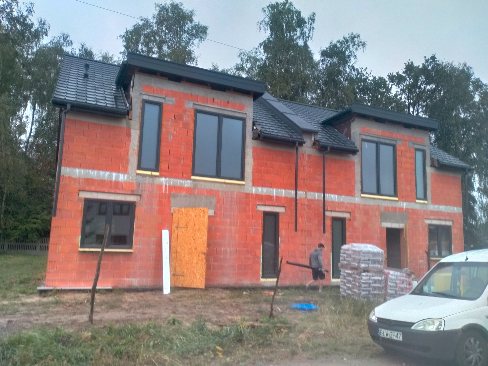 Surowy, dwukondygnacyjny dom z czerwonej cegły w trakcie budowy. Widoczne czarne okna, szara dachówka, materiały budowlane i częściowo zakryte wejście.