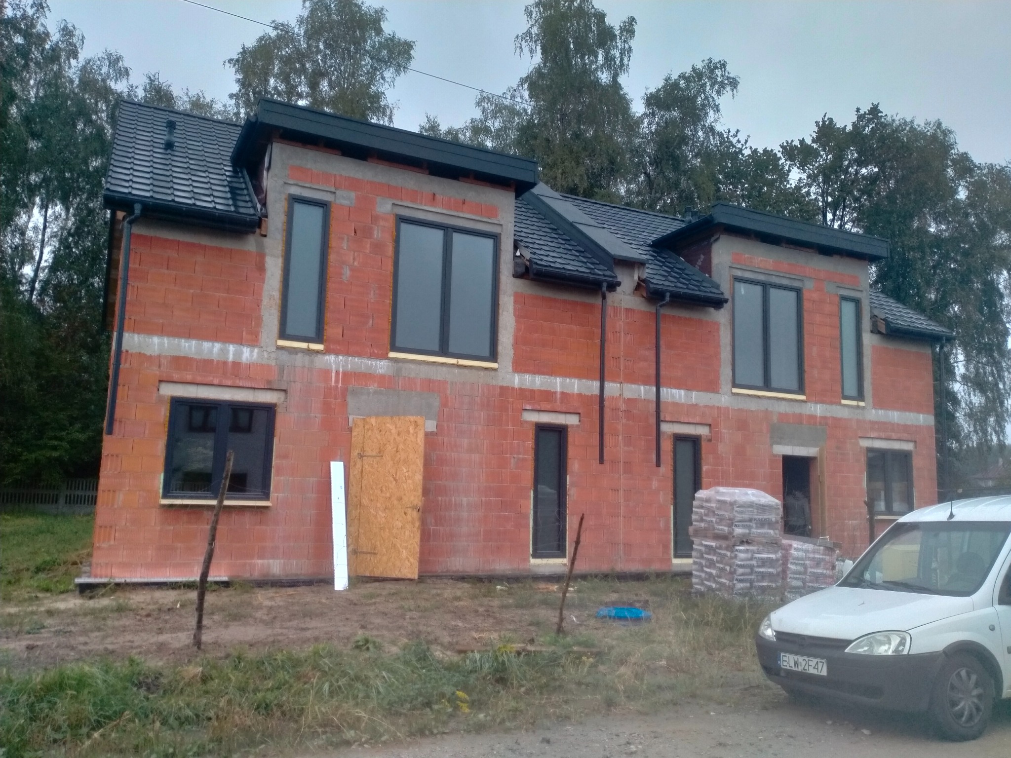 Surowy stan dwukondygnacyjnego domu z czerwonej cegły z czarną dachówką i oknami, widoczne materiały budowlane i samochód dostawczy na zewnątrz.
