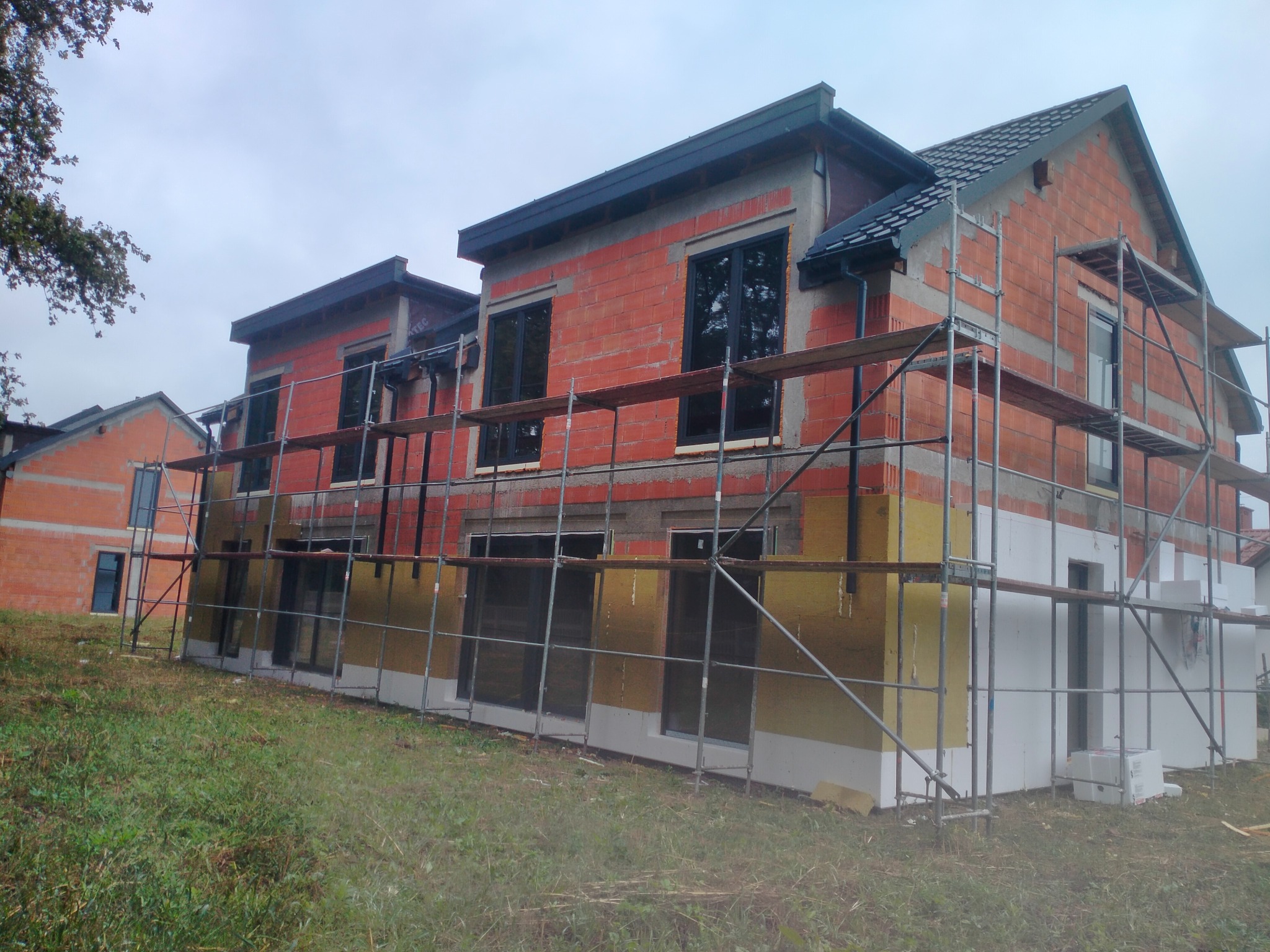 Budowa domu z czerwonej cegły, częściowo ocieplonego, z rusztowaniami. Widoczne okna i dach. Trawa na pierwszym planie, pochmurne niebo w tle. Ujęcie z dołu.